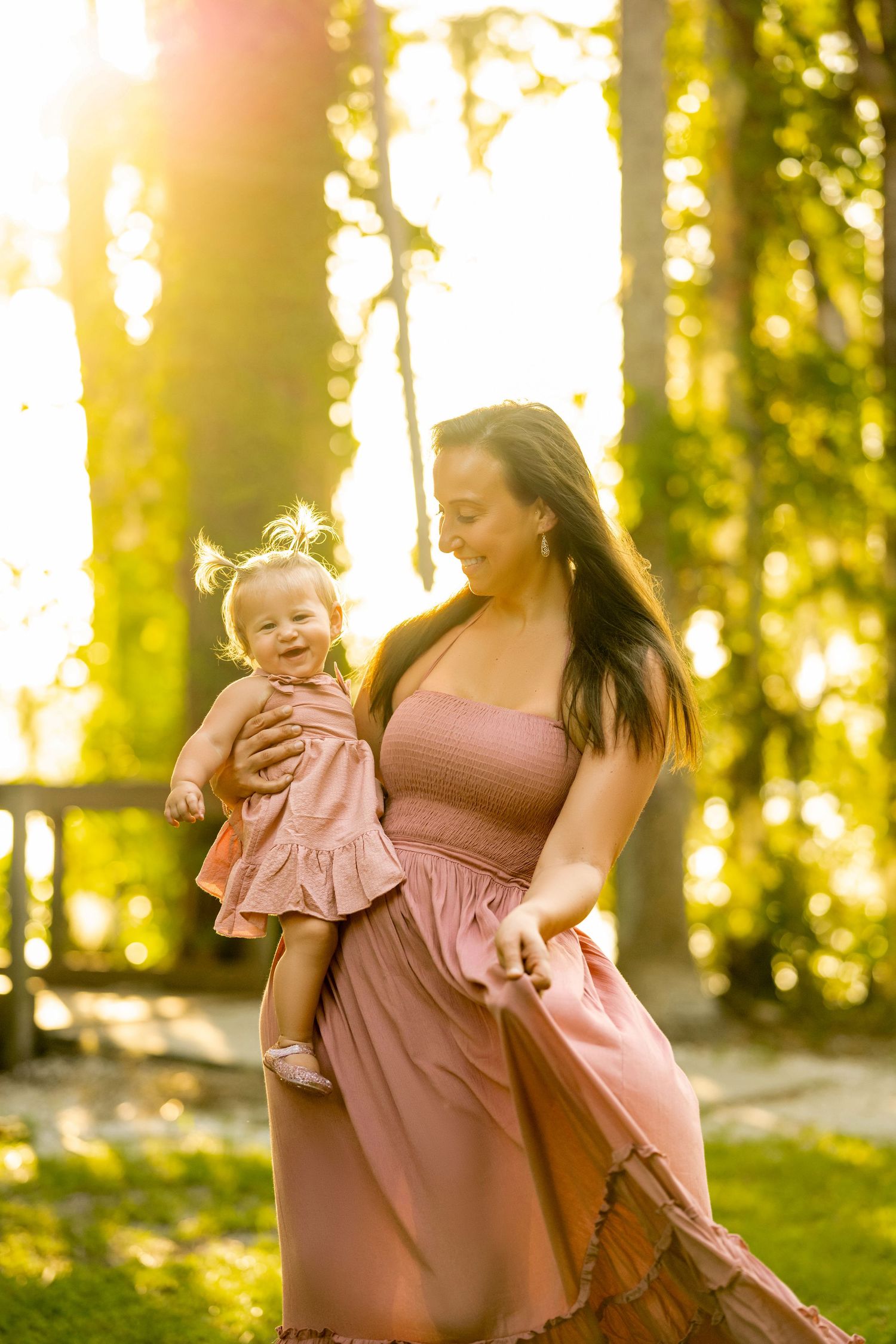 Mother in pink dress holds smiling baby girl outdoors in golden sunlight among green trees.