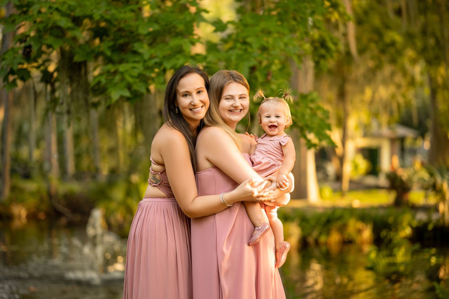Two women in pink dresses smile holding a baby girl outdoors near a pond with lush green trees.