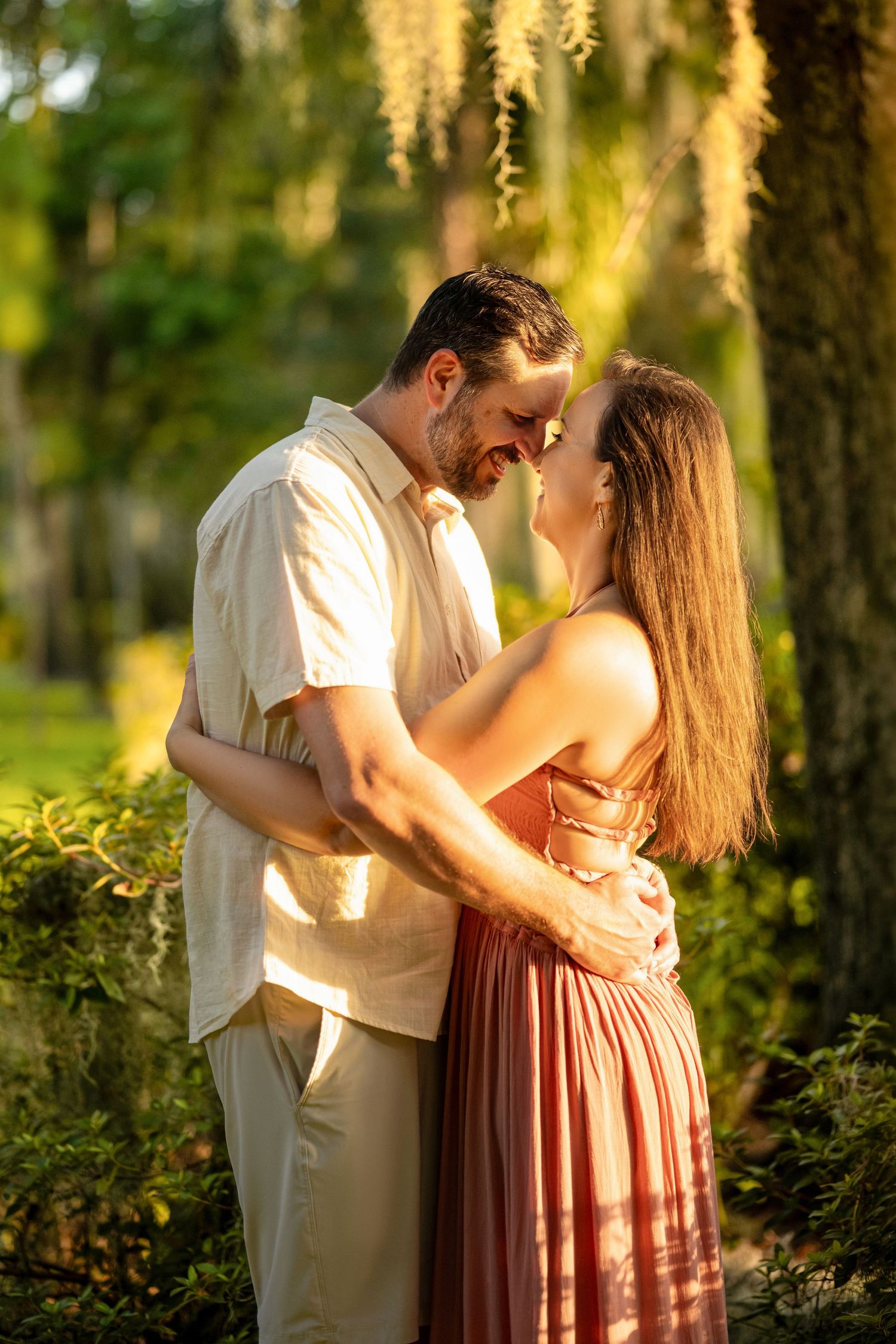 Couple embracing intimately in a sunlit park with Spanish moss hanging from trees at golden hour.