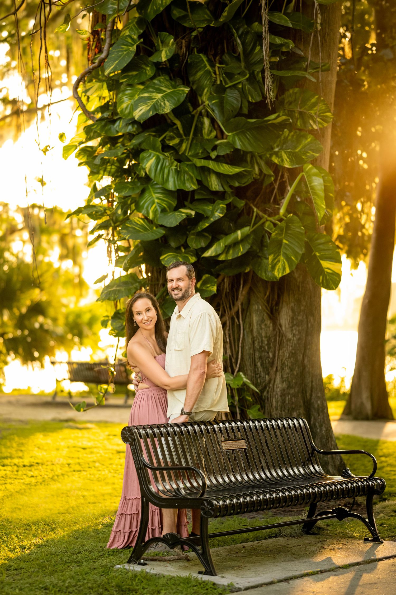 Romantic couple hugging beside a park bench under a large tree with lush foliage at sunset.