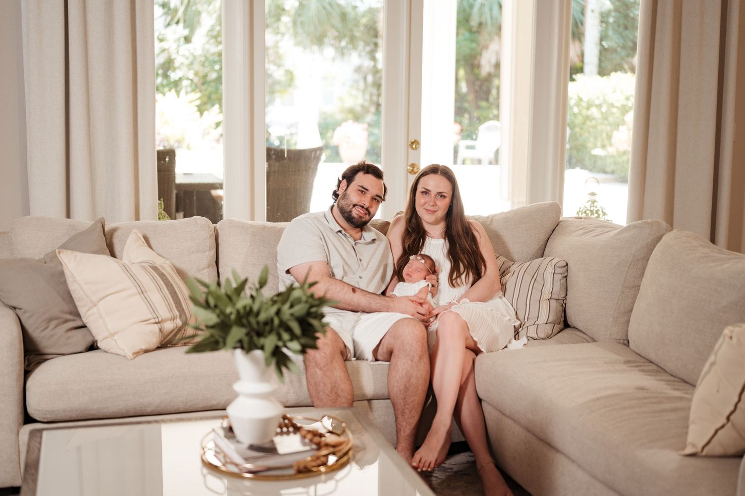 A young family with a newborn baby poses on a beige sectional sofa in a bright, modern living room.
