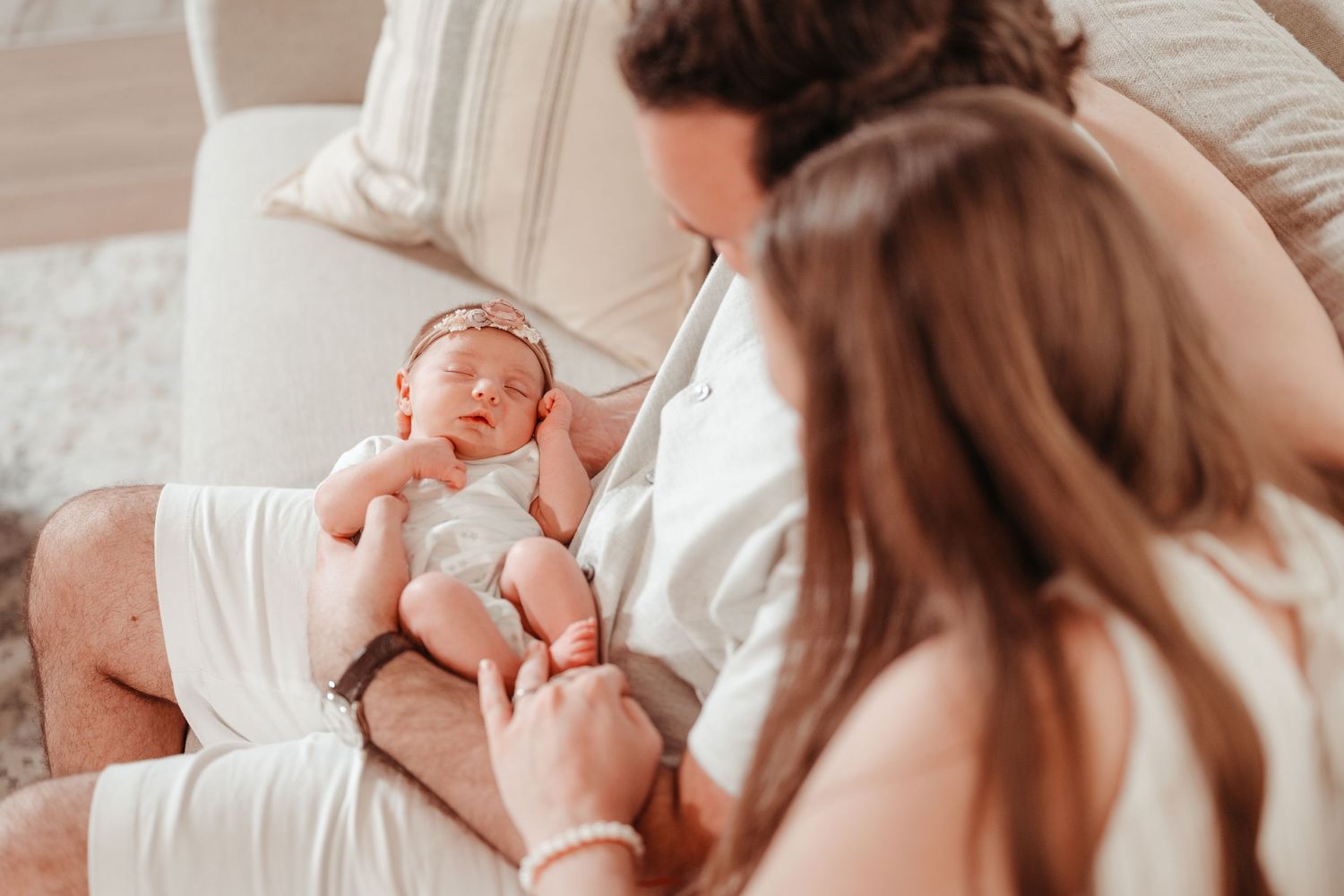 Parents admiring their sleeping newborn baby wrapped in white, resting on father's lap during an intimate family moment.