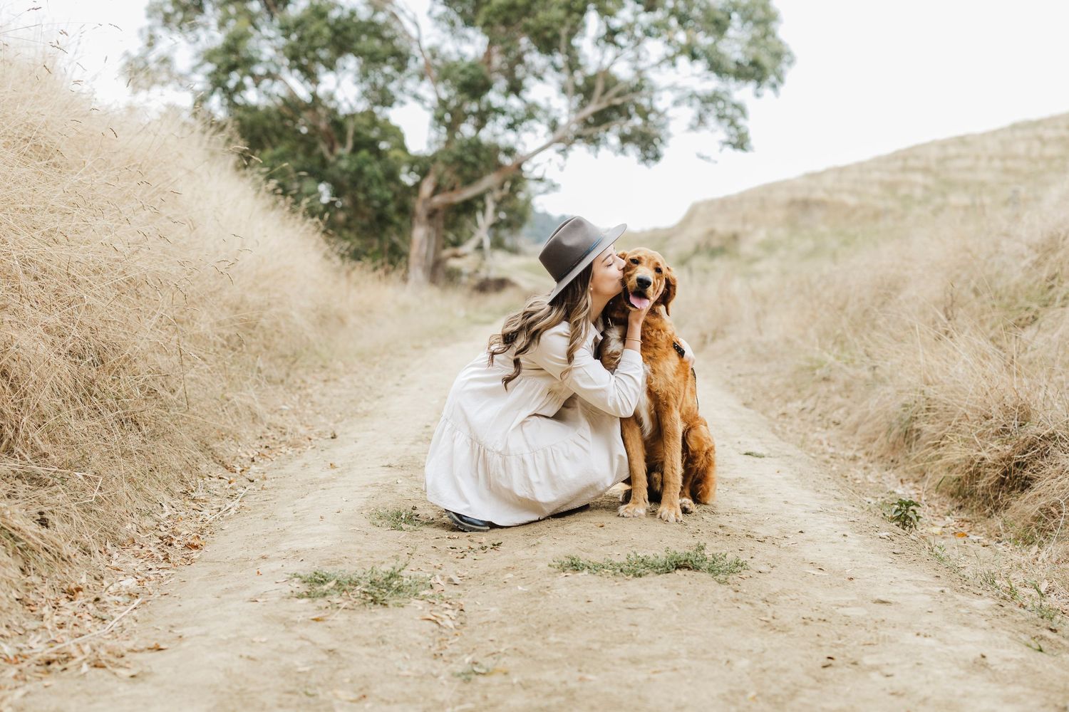 A person in a flowing white dress sits on a dirt path with their golden retriever companion in a rustic outdoor setting.