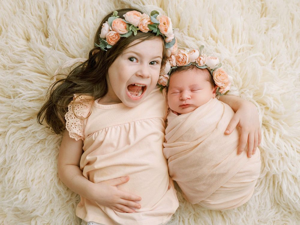 Two children wearing peach colored outfits and floral crowns lay together on white fluffy fabric.