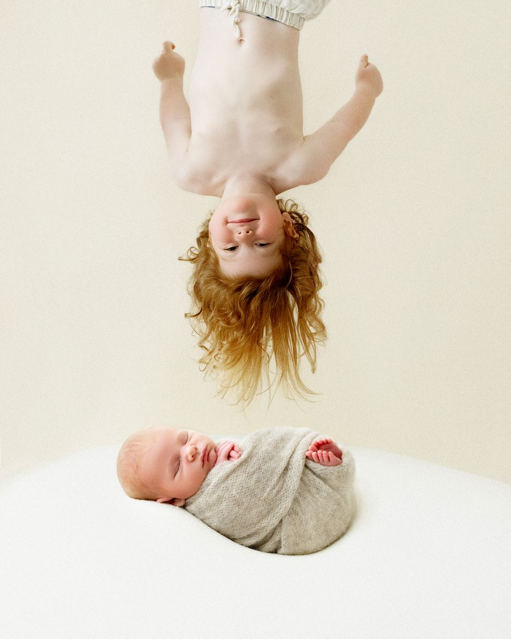 Creative photo of one child suspended upside down above another laying on white surface in gray swaddle.