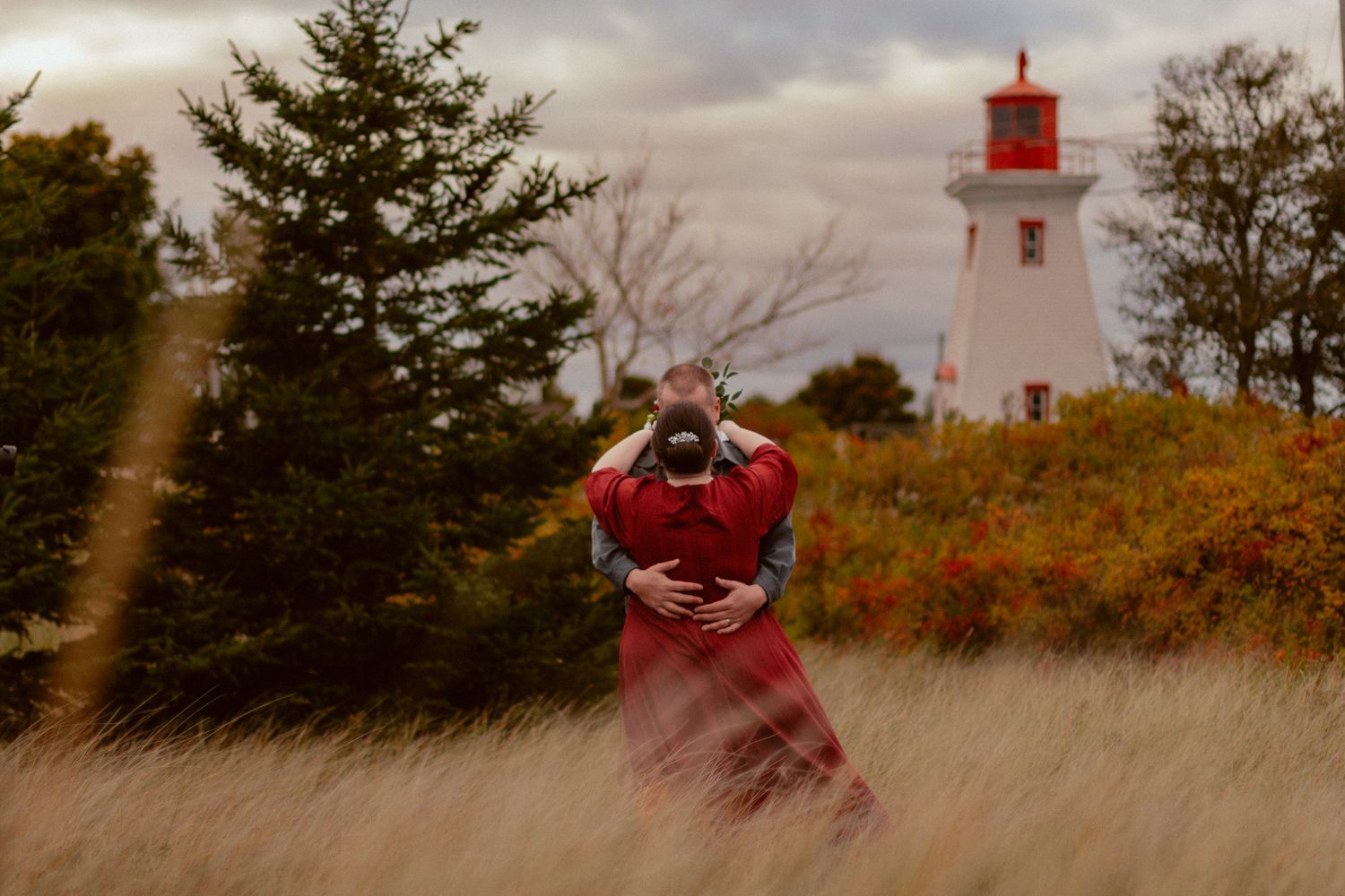 A red dress stands out against tall grass with a lighthouse in the background.