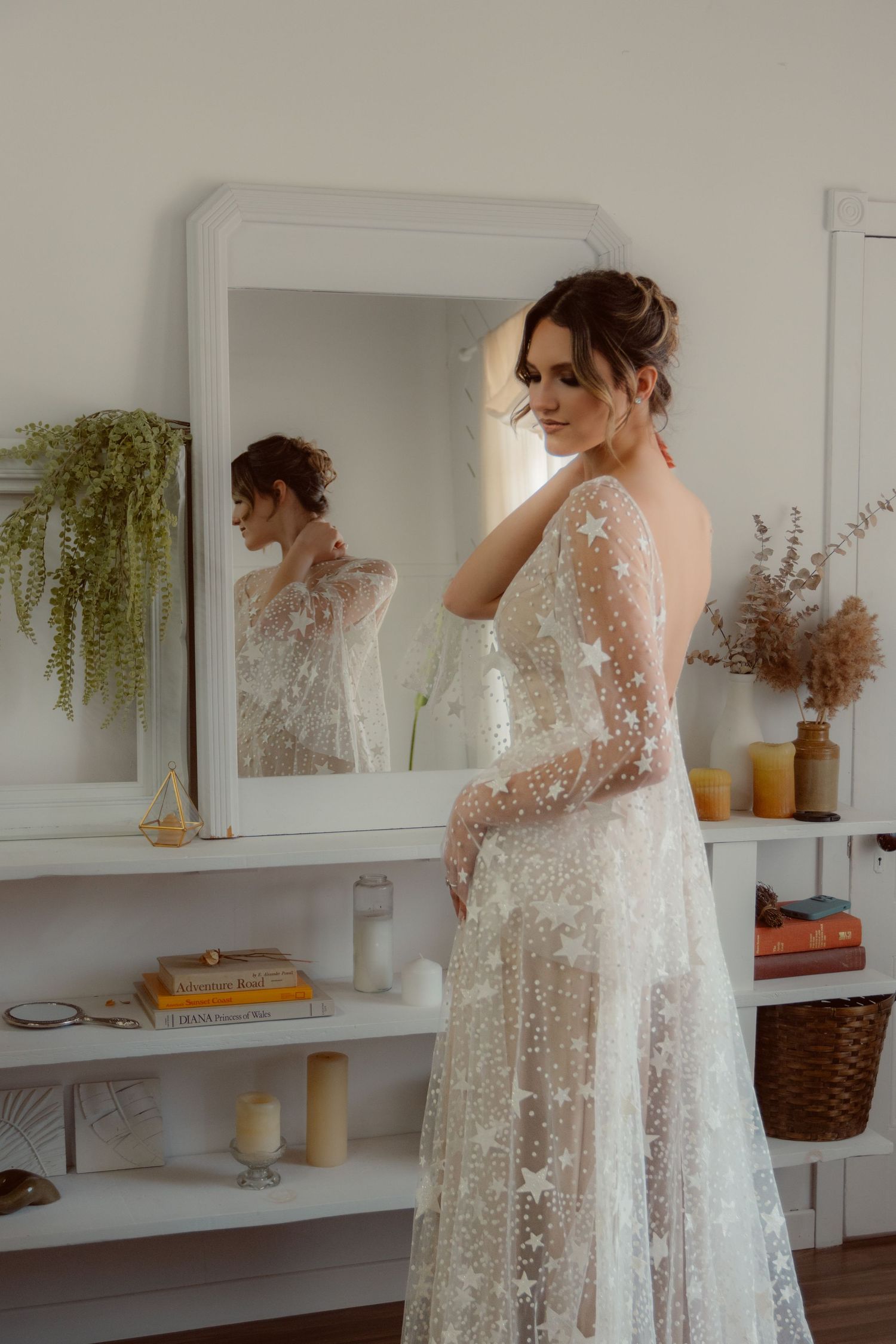 Bride in lace wedding dress looking at her reflection in elegant white room with shelving.