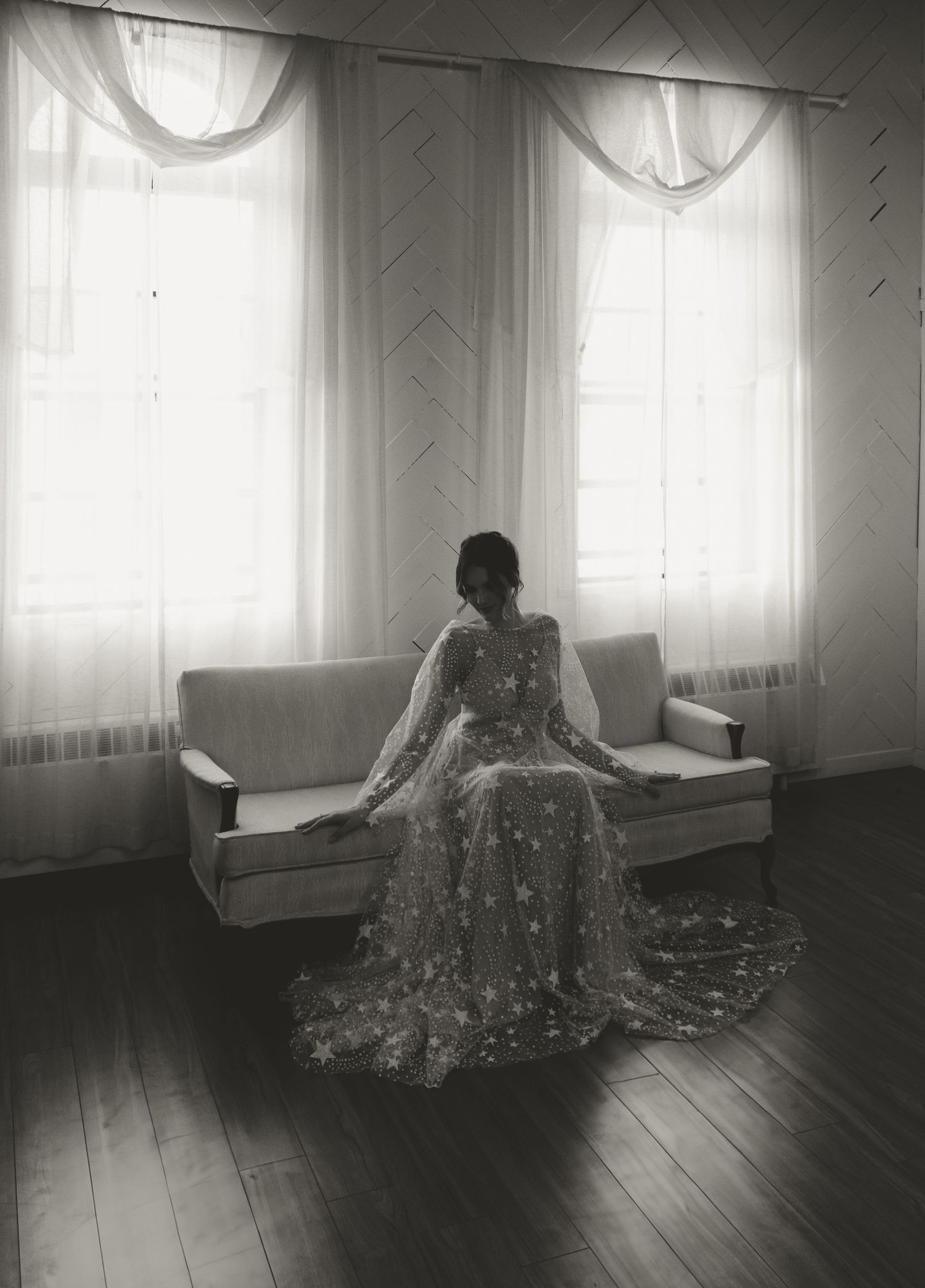 Vintage-style photo of bride in lace dress seated by bright windows with sheer curtains.