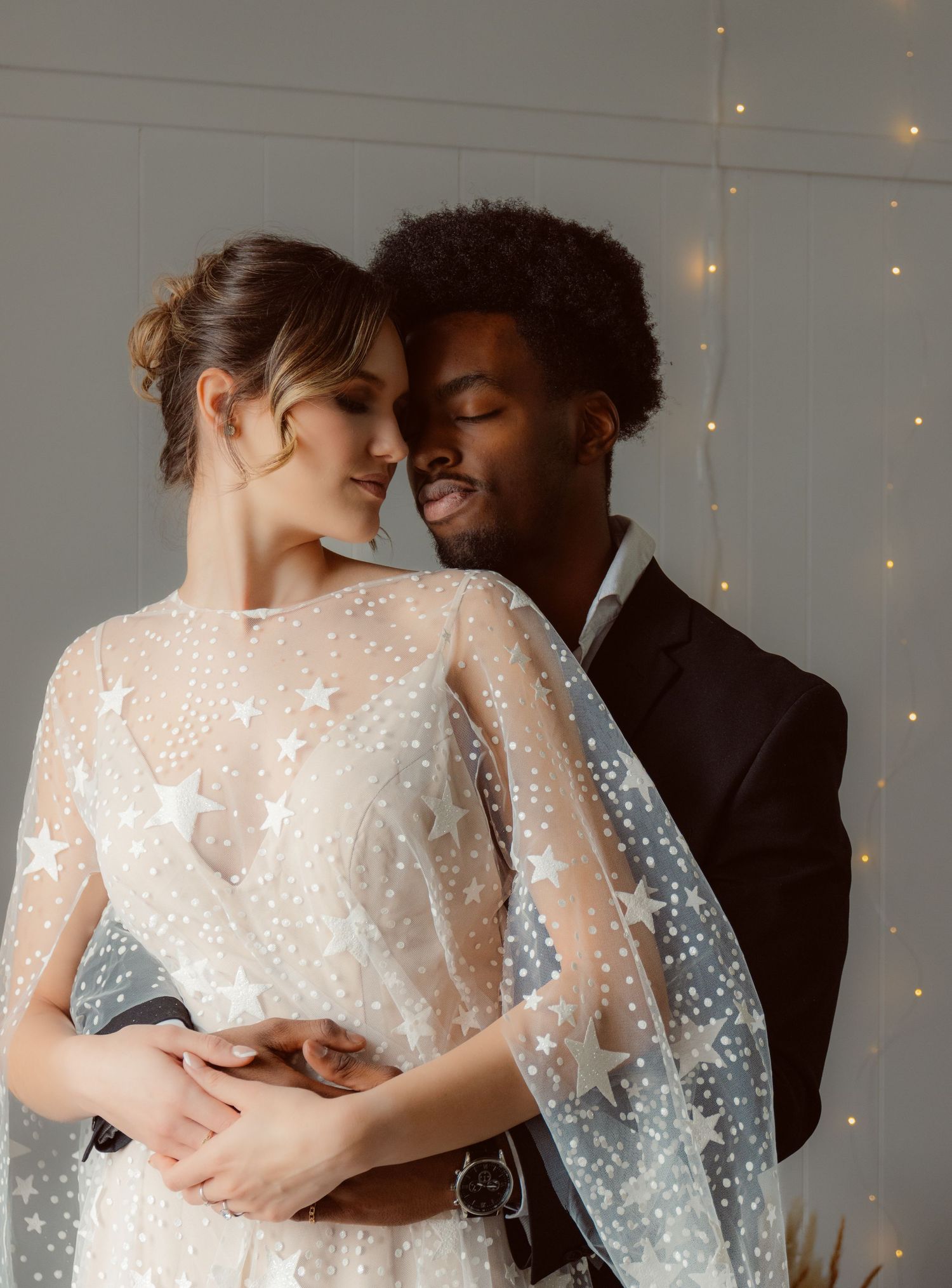 Romantic wedding portrait of couple in embrace against white wall with string lights.