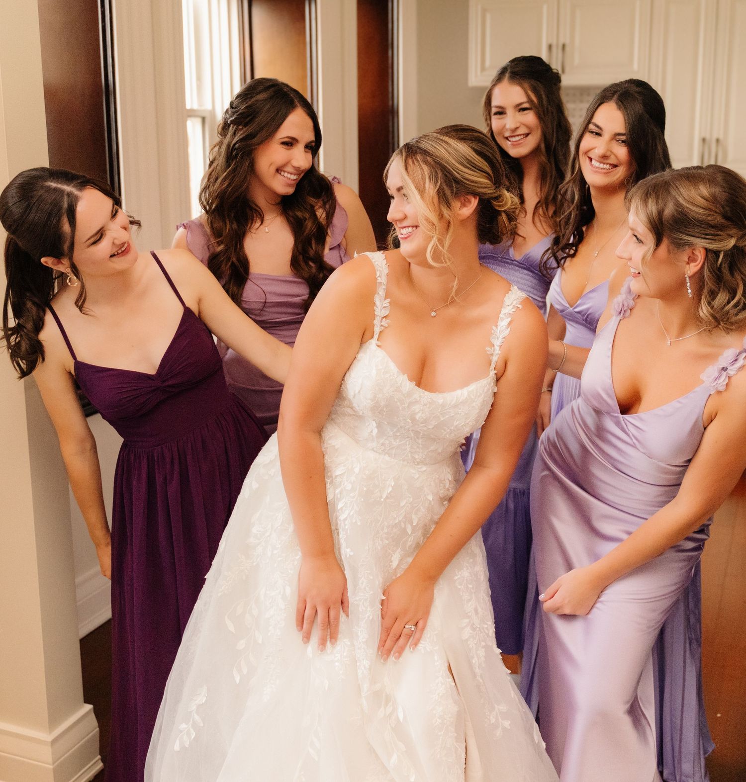 Bridesmaids in purple dresses helping with final touches on bride's wedding gown.