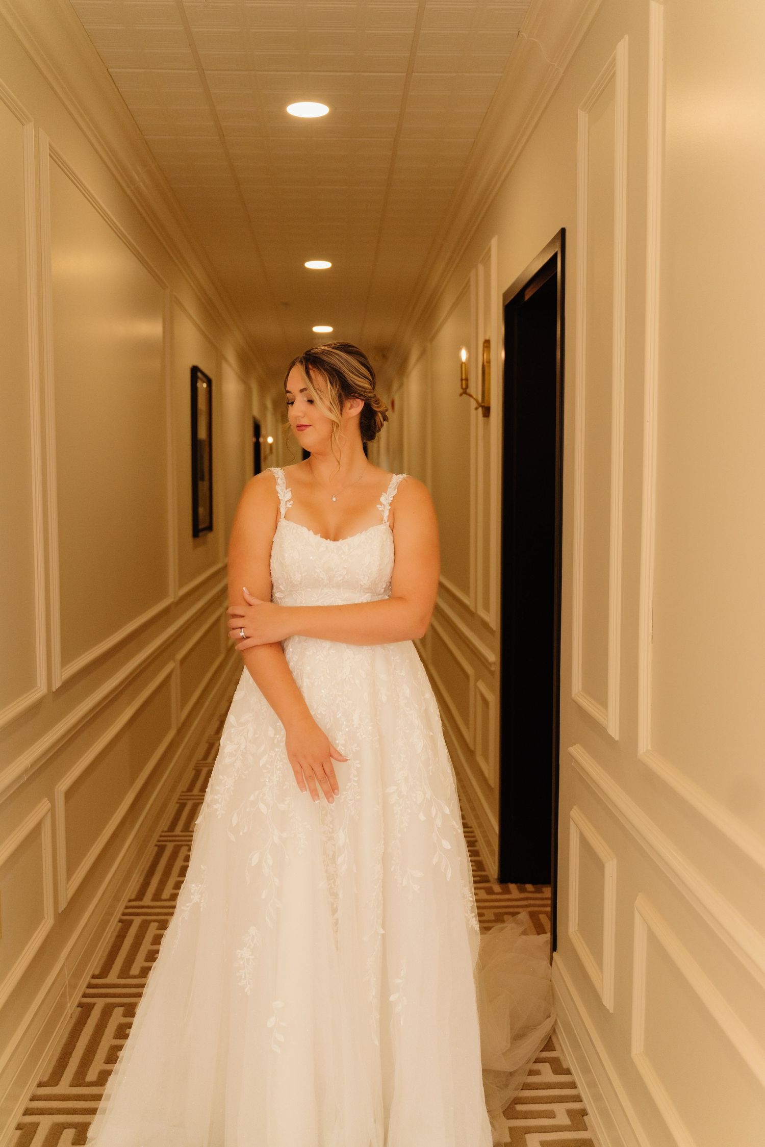 Bride in white lace gown standing alone in elegant hotel hallway.