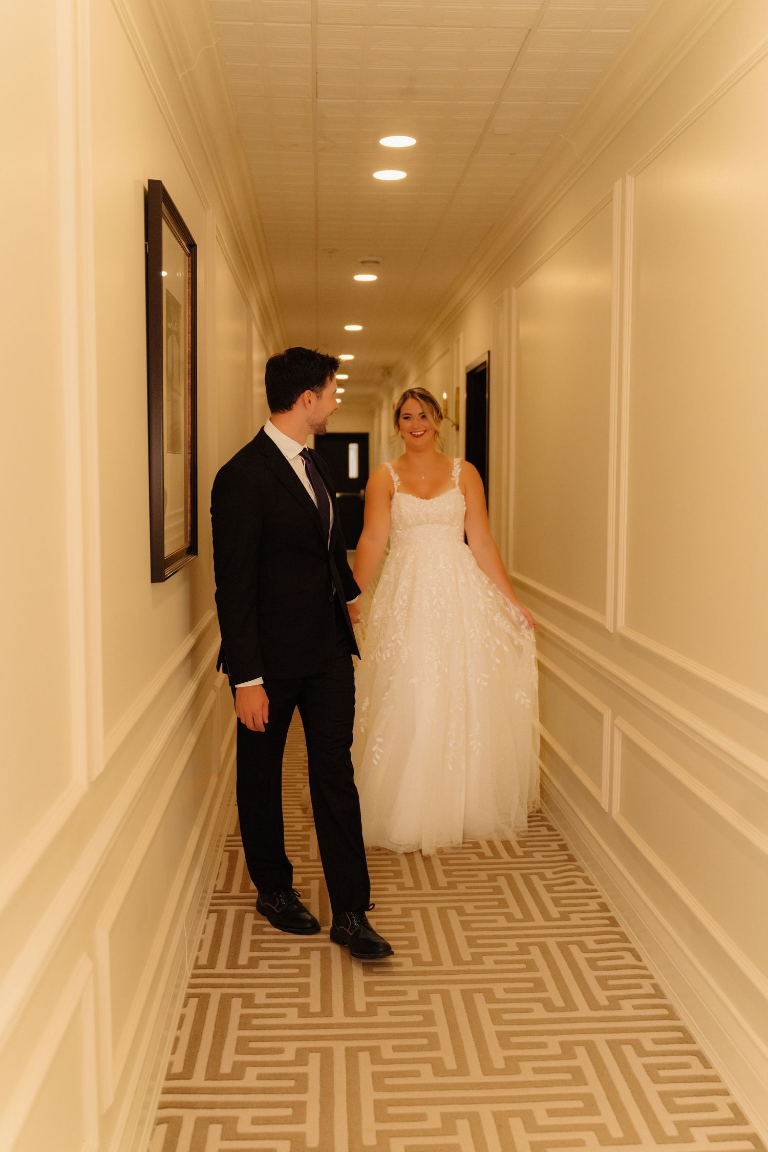 Couple in wedding attire meeting in elegant hotel hallway with geometric carpet.