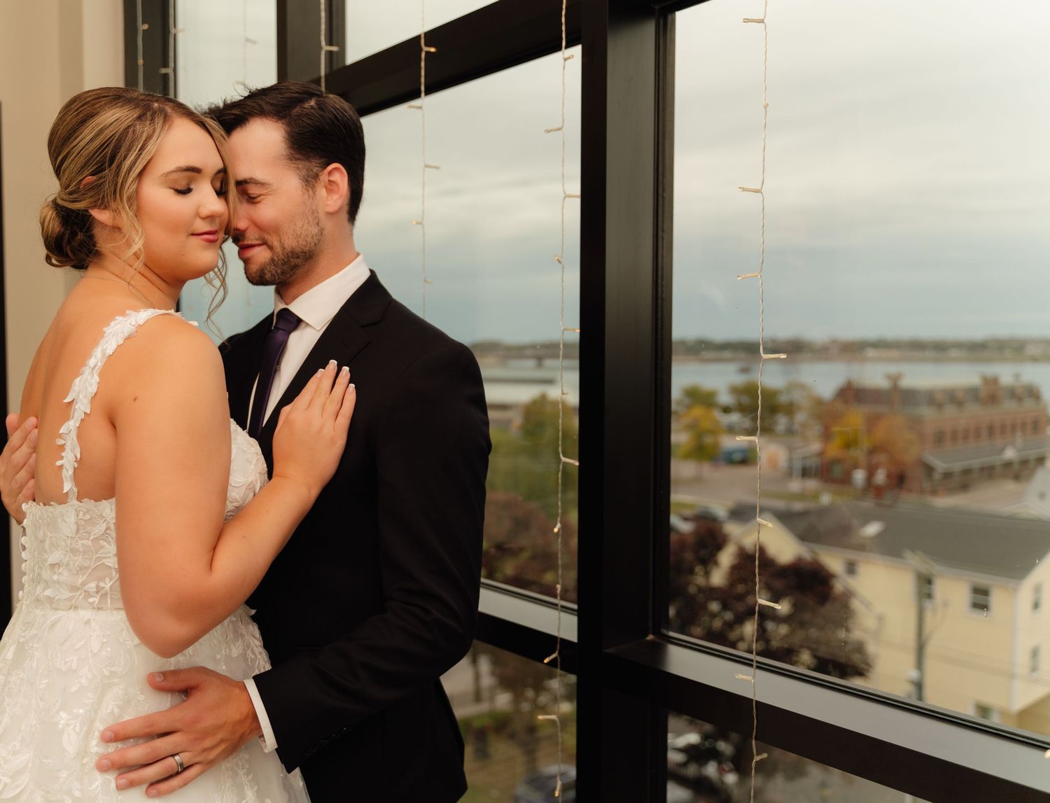 Intimate moment between newlyweds by window overlooking city view.