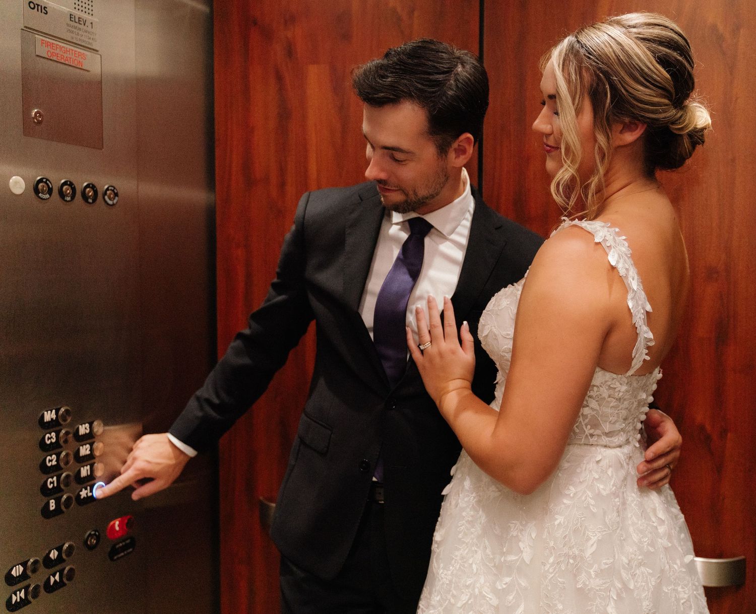 A couple in wedding attire press the elevator button together.