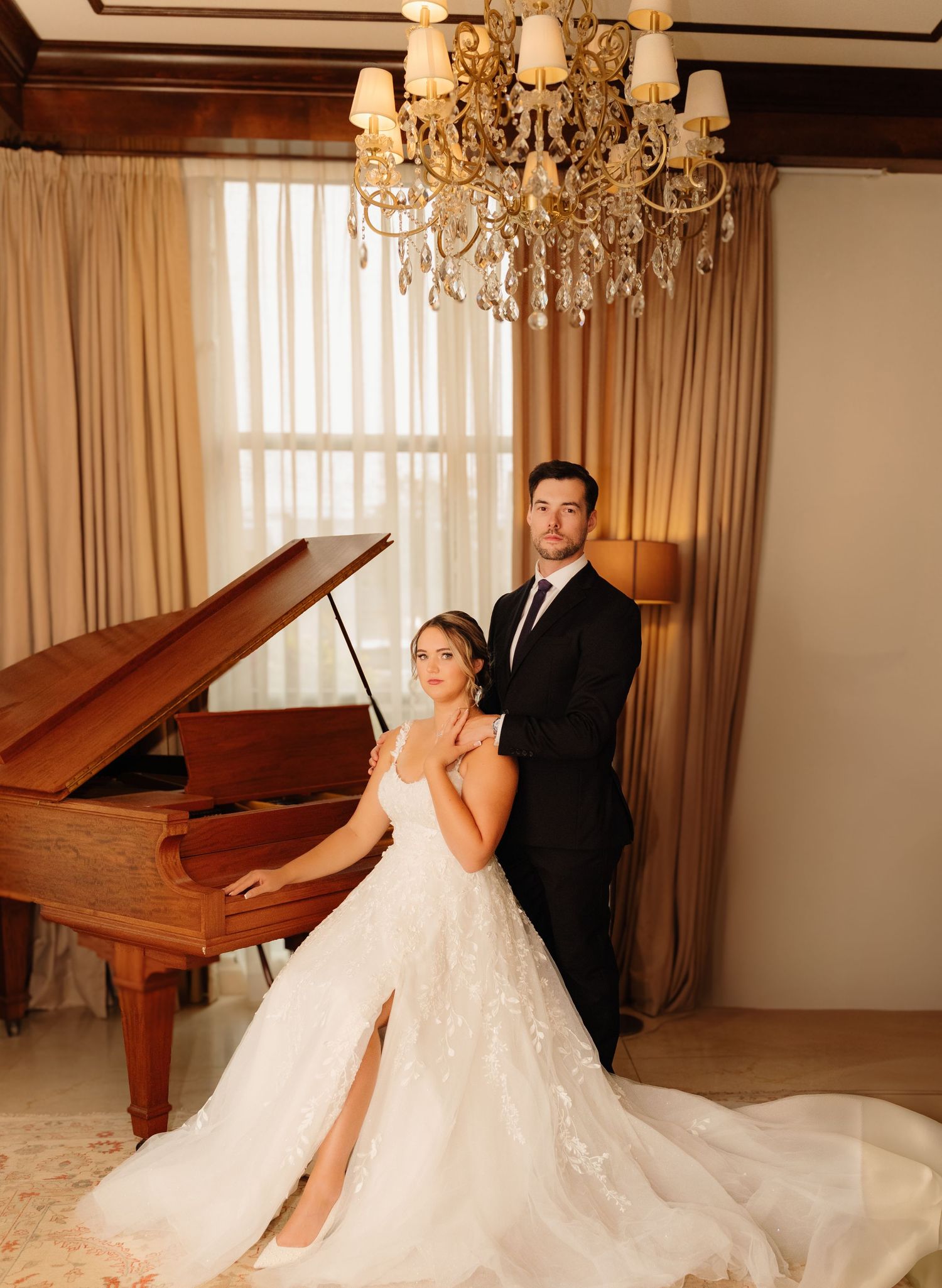 A wedding couple poses by a grand piano under an elegant crystal chandelier.