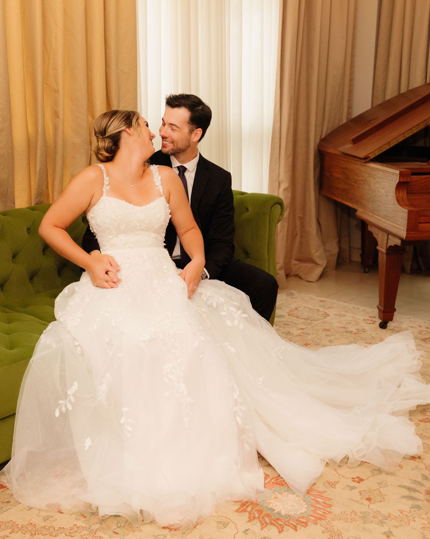 Newlyweds share an intimate moment on a green velvet couch near a piano.