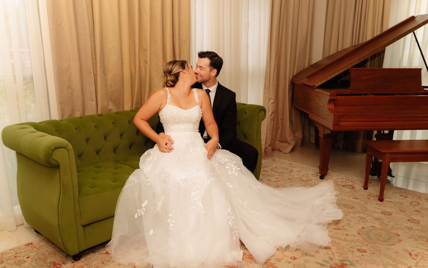 A couple in wedding attire sits together on a green velvet sofa beside a grand piano.