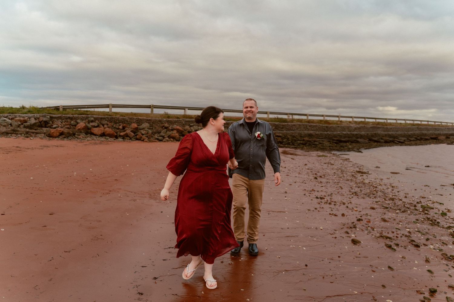 A couple walks together on a beach with rust-colored sand and cloudy skies.