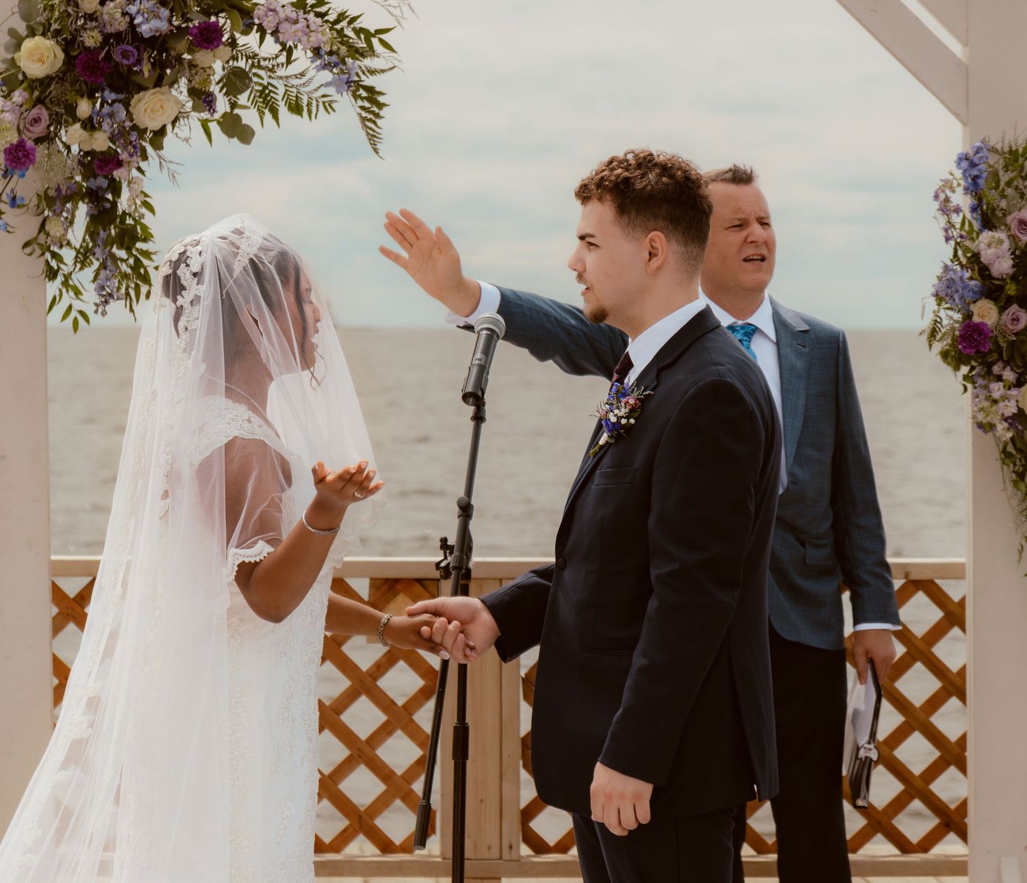 A wedding ceremony takes place under a floral arch by the waterfront.