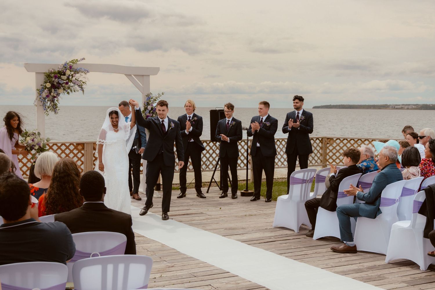 Wedding party and guests gather on a waterfront deck during an outdoor ceremony.