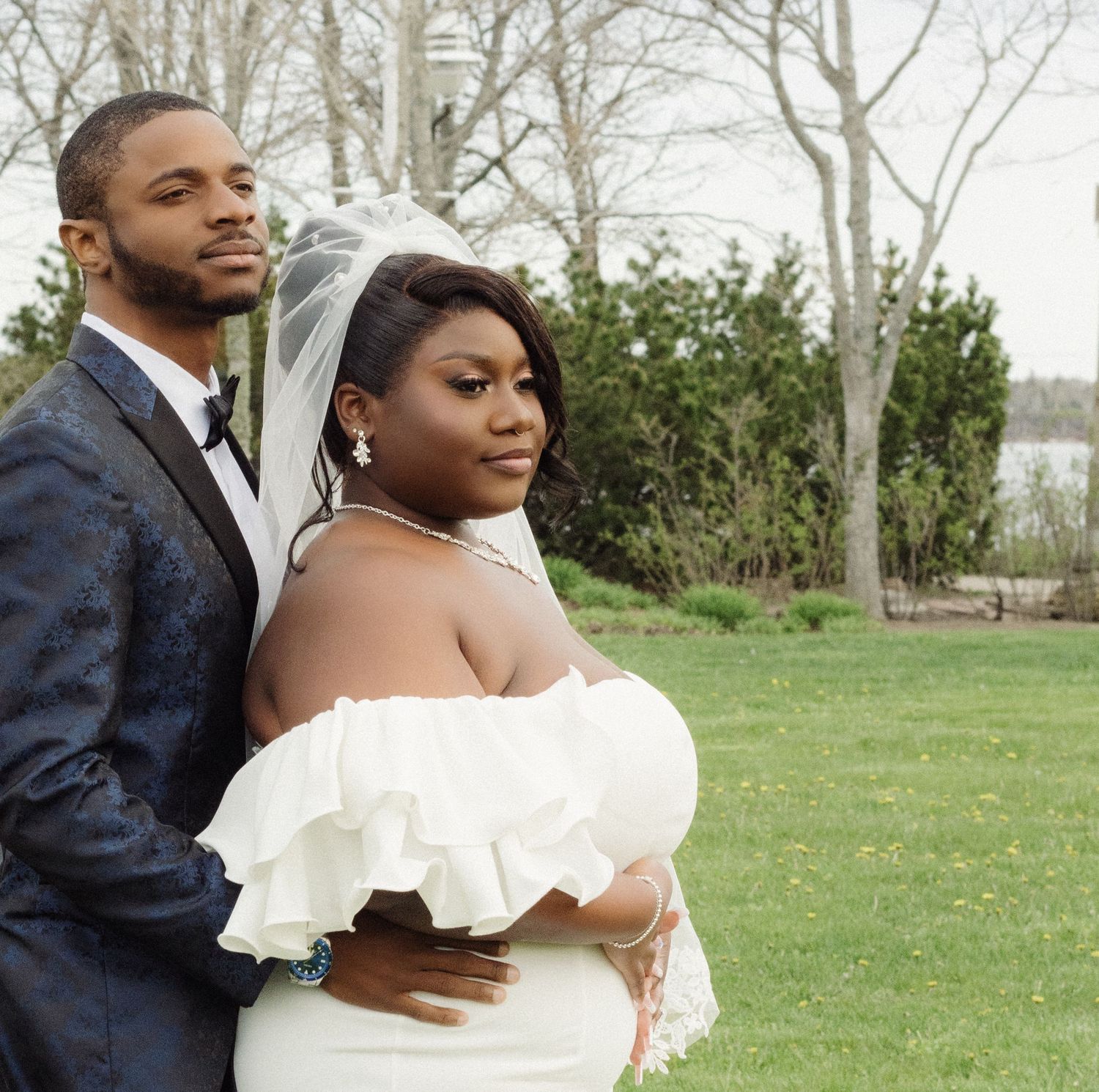 An elegant wedding couple poses together on a green lawn with bare trees in the background.