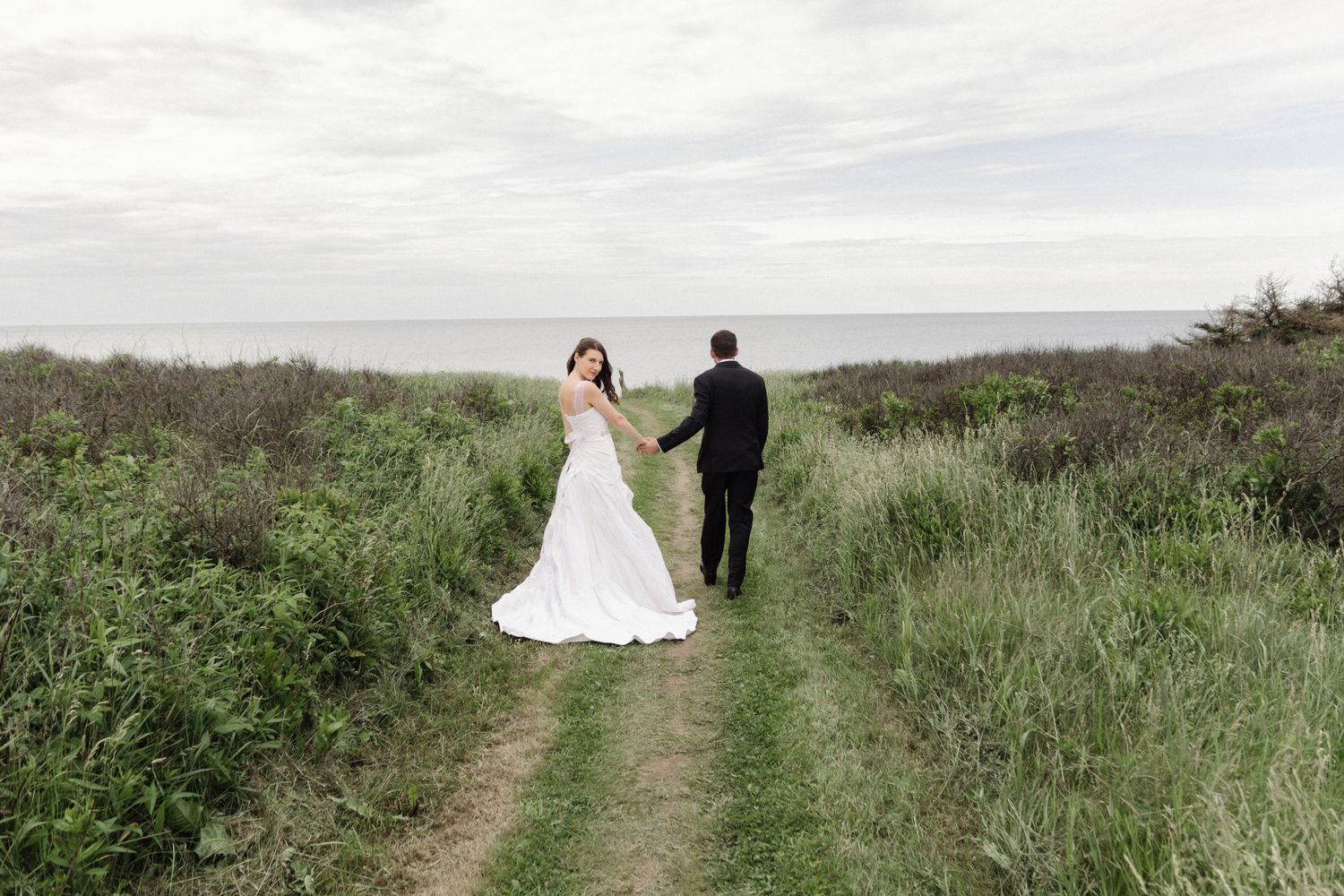 A newlywed couple walks down a grassy path surrounded by natural landscape.