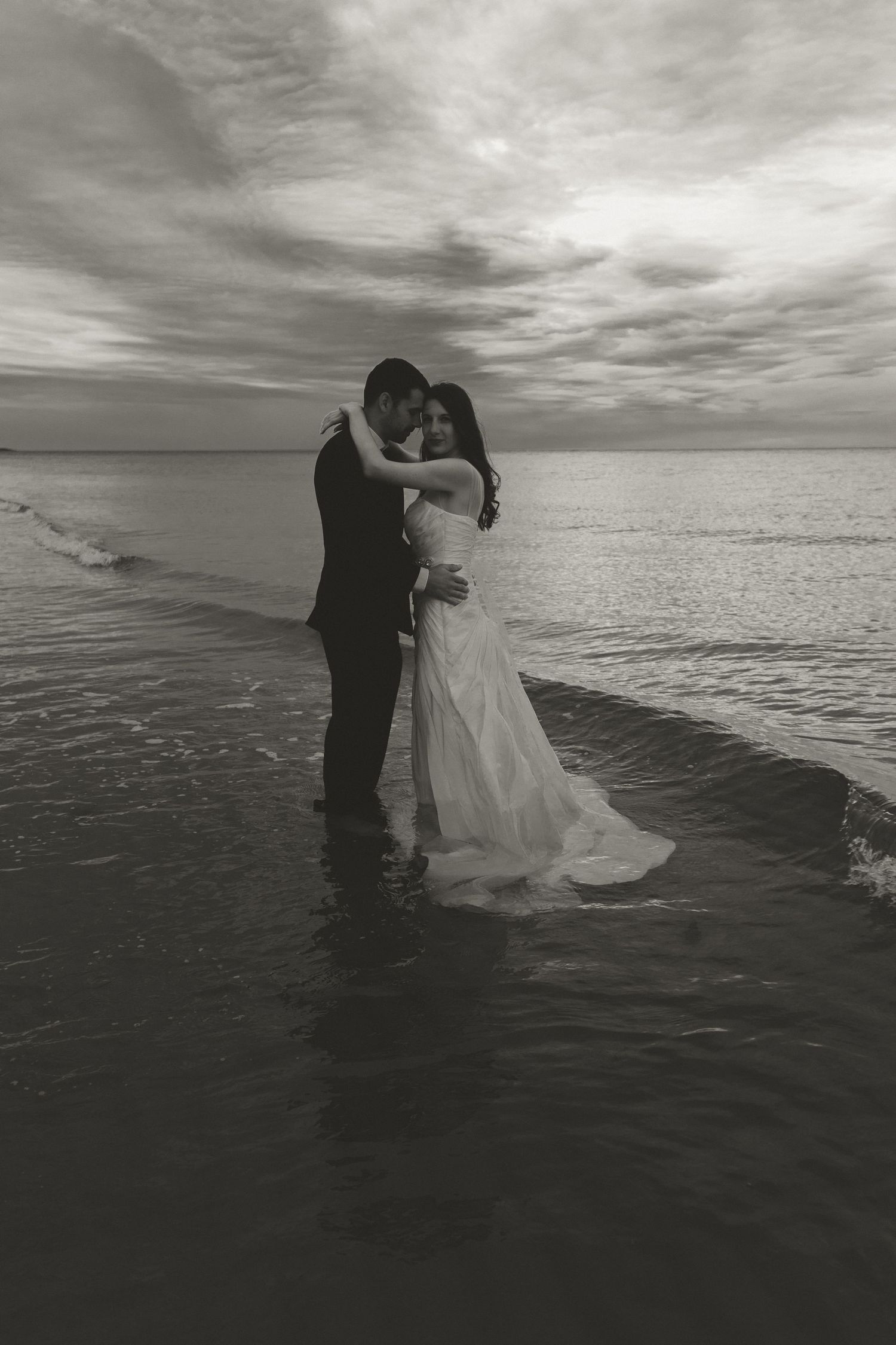 A black and white photo of newlyweds embracing on a beach at sunset with dramatic cloudy skies.