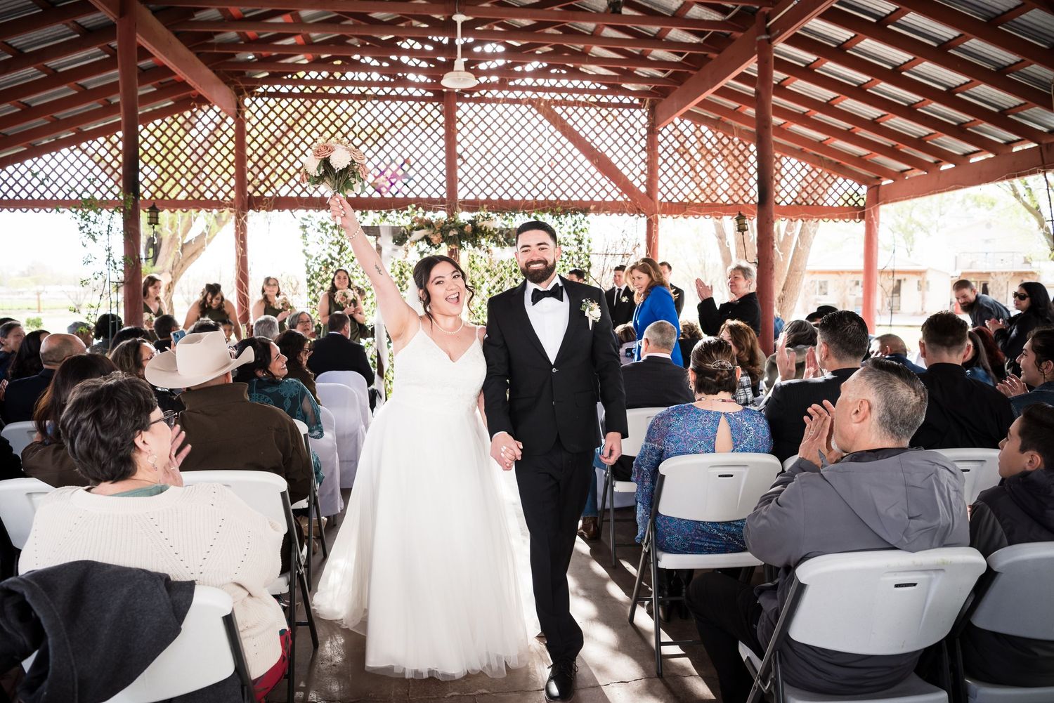 A wedding ceremony procession in a rustic covered pavilion filled with guests.