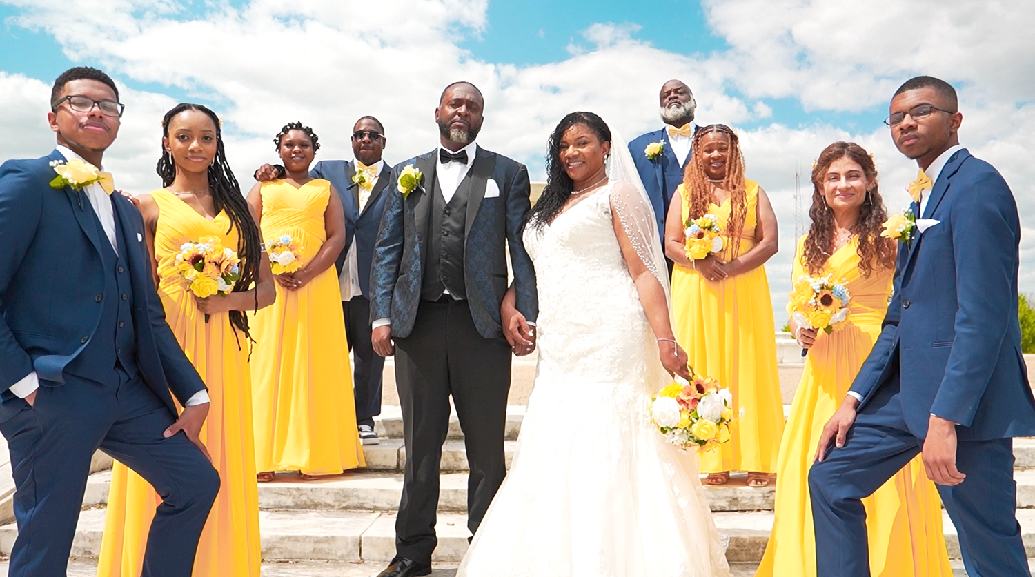 Wedding party in yellow bridesmaid dresses and navy suits pose outdoors against a bright blue sky.