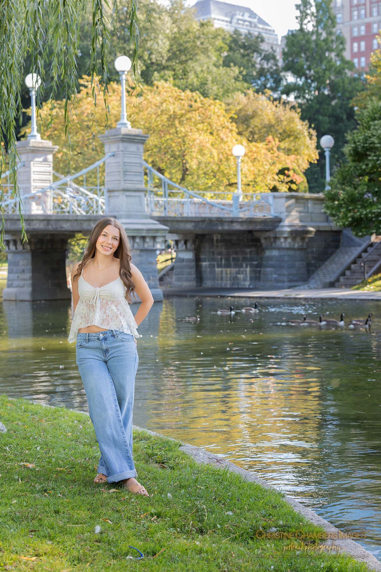 A scenic pond in Boston Public Garden with an ornate stone bridge and golden autumn foliage in the background. High school senior girl