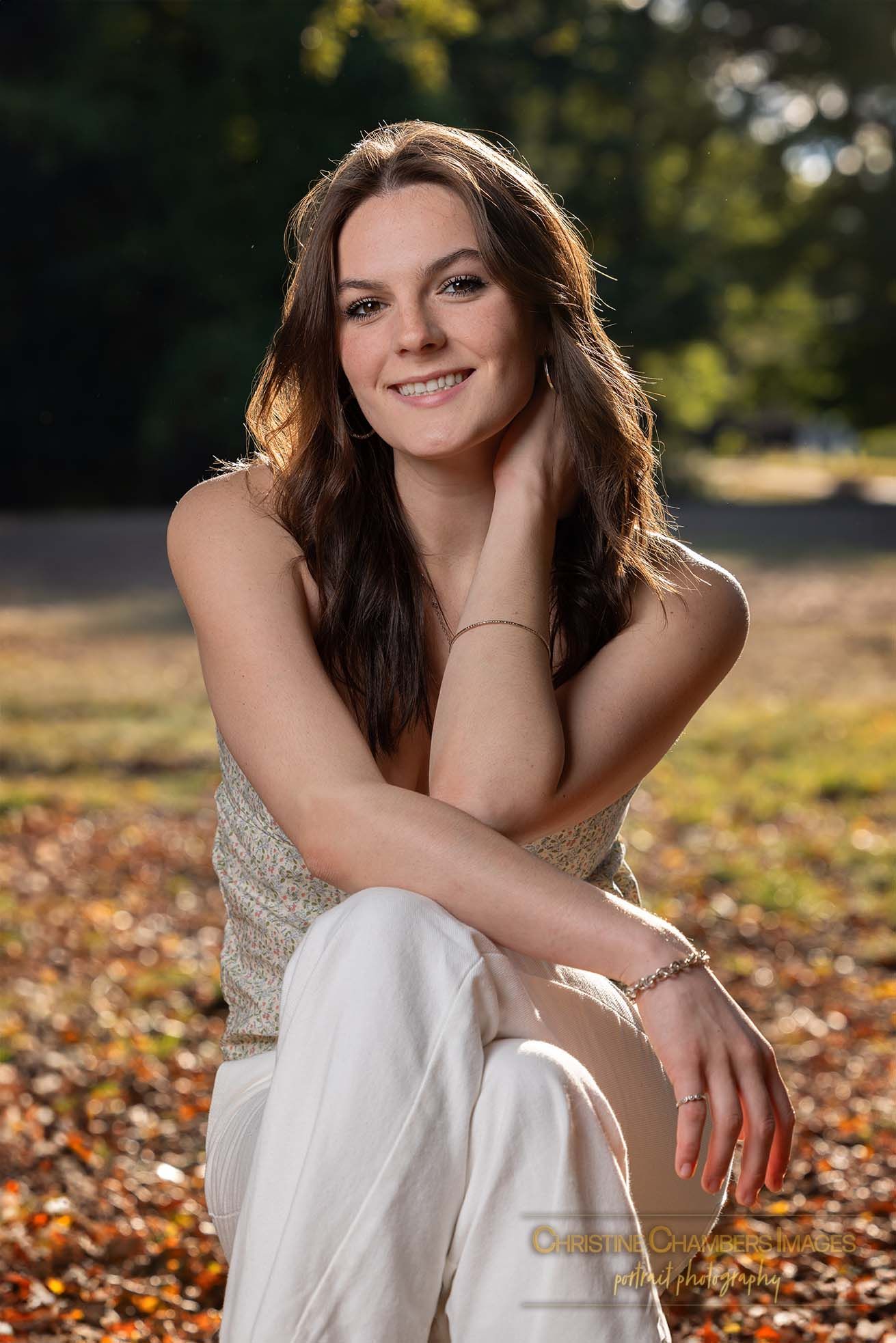 A person in a white dress poses amid autumn leaves while sitting outdoors in golden sunlight.  High school senior girl