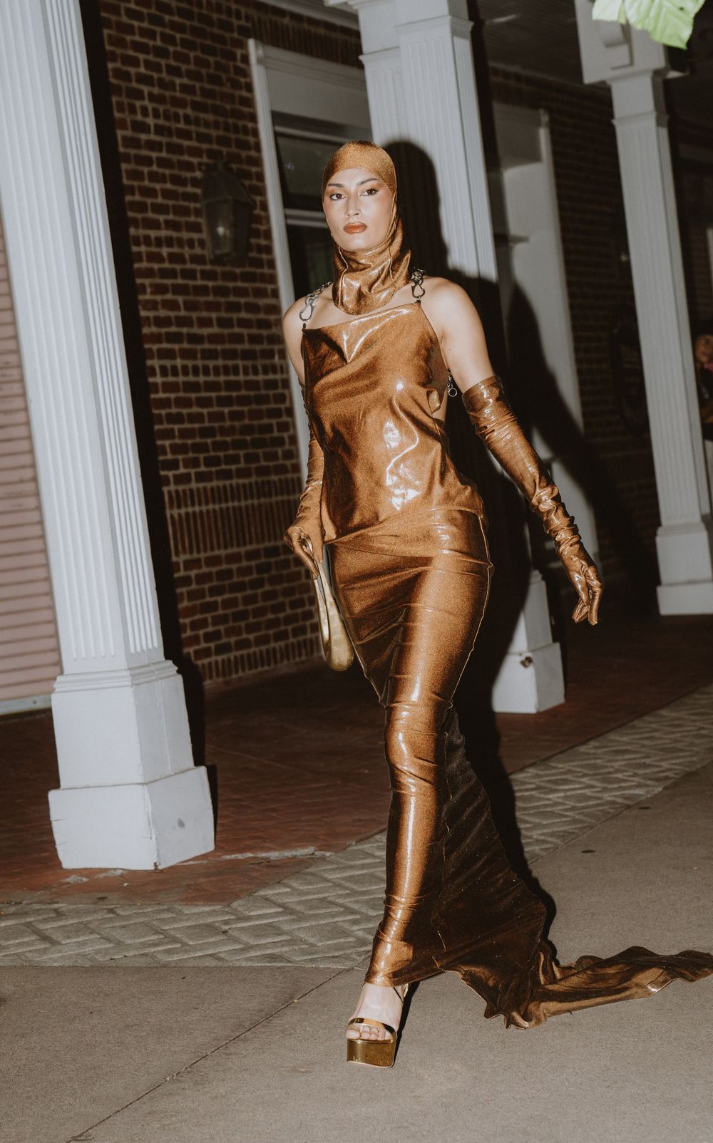 Fashion model walks in metallic copper bodysuit and gloves against brick wall.