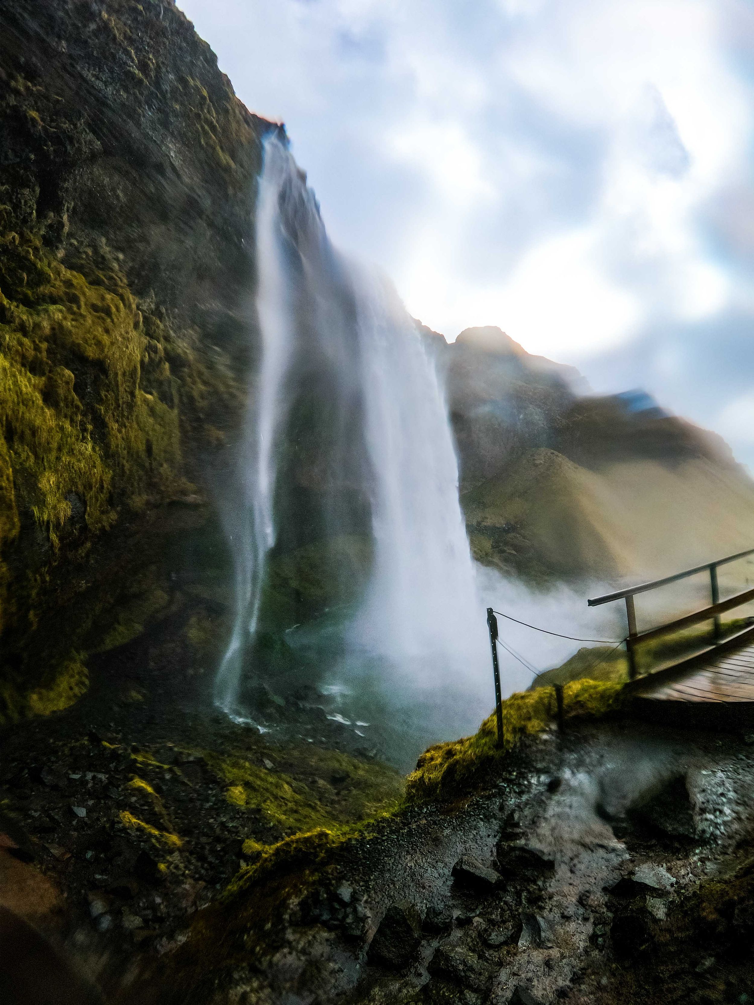 Towering waterfall cascading down moss-covered cliffs with viewing bridge in foreground.