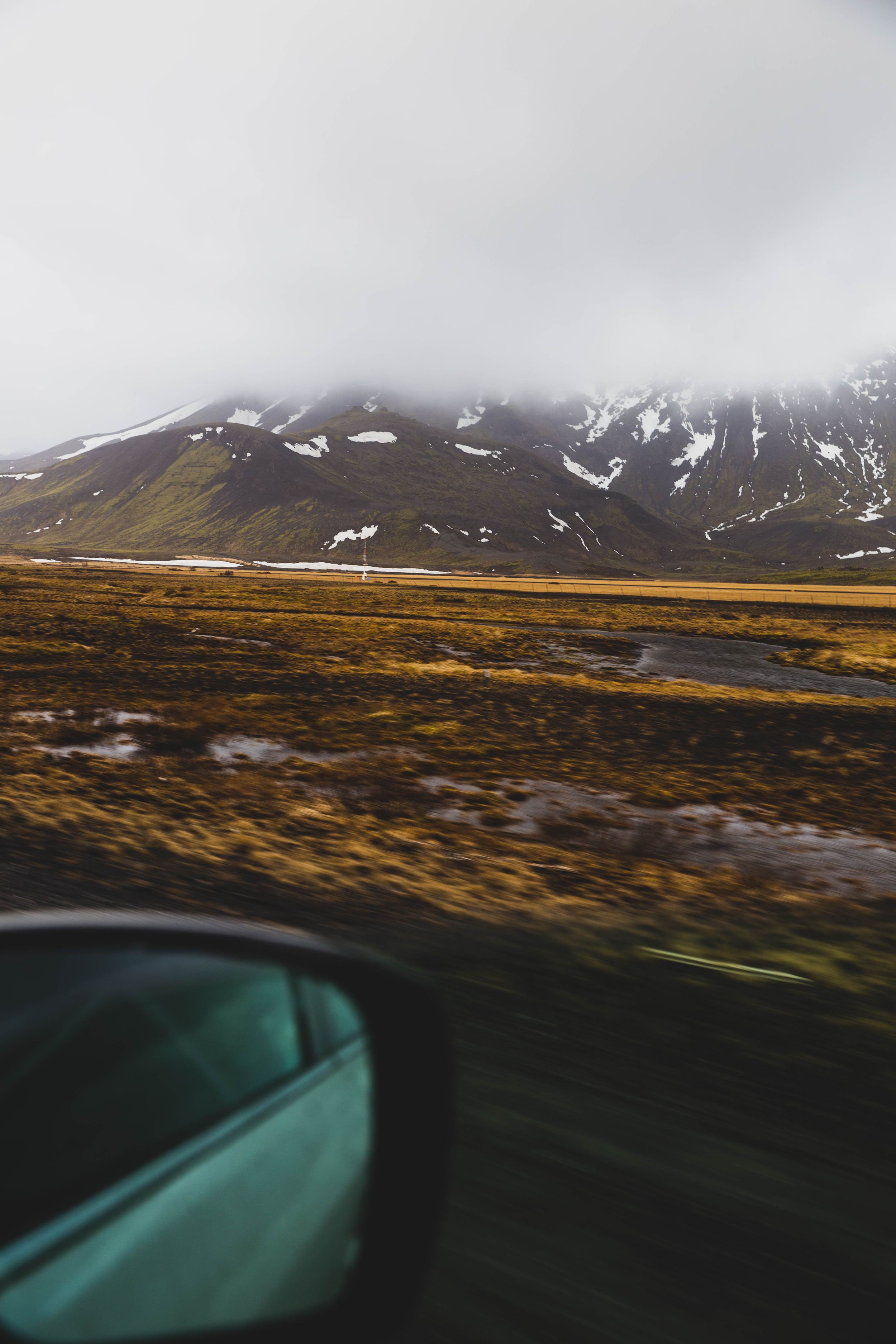 Snow-capped mountains and golden tundra landscape viewed through a car side mirror on a cloudy day.