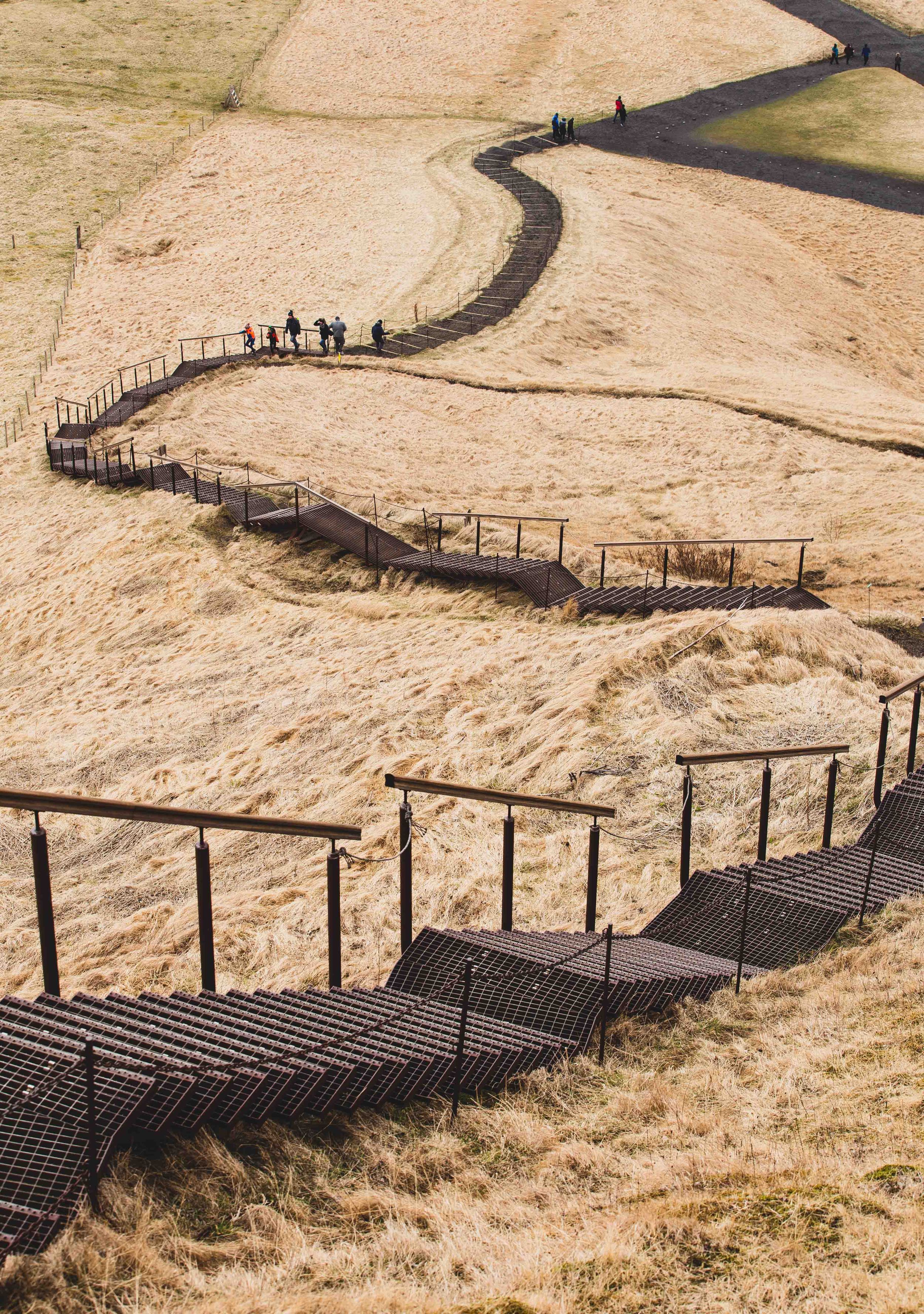 Winding wooden boardwalk path curves through dry grassland with distant hikers visible along the trail.
