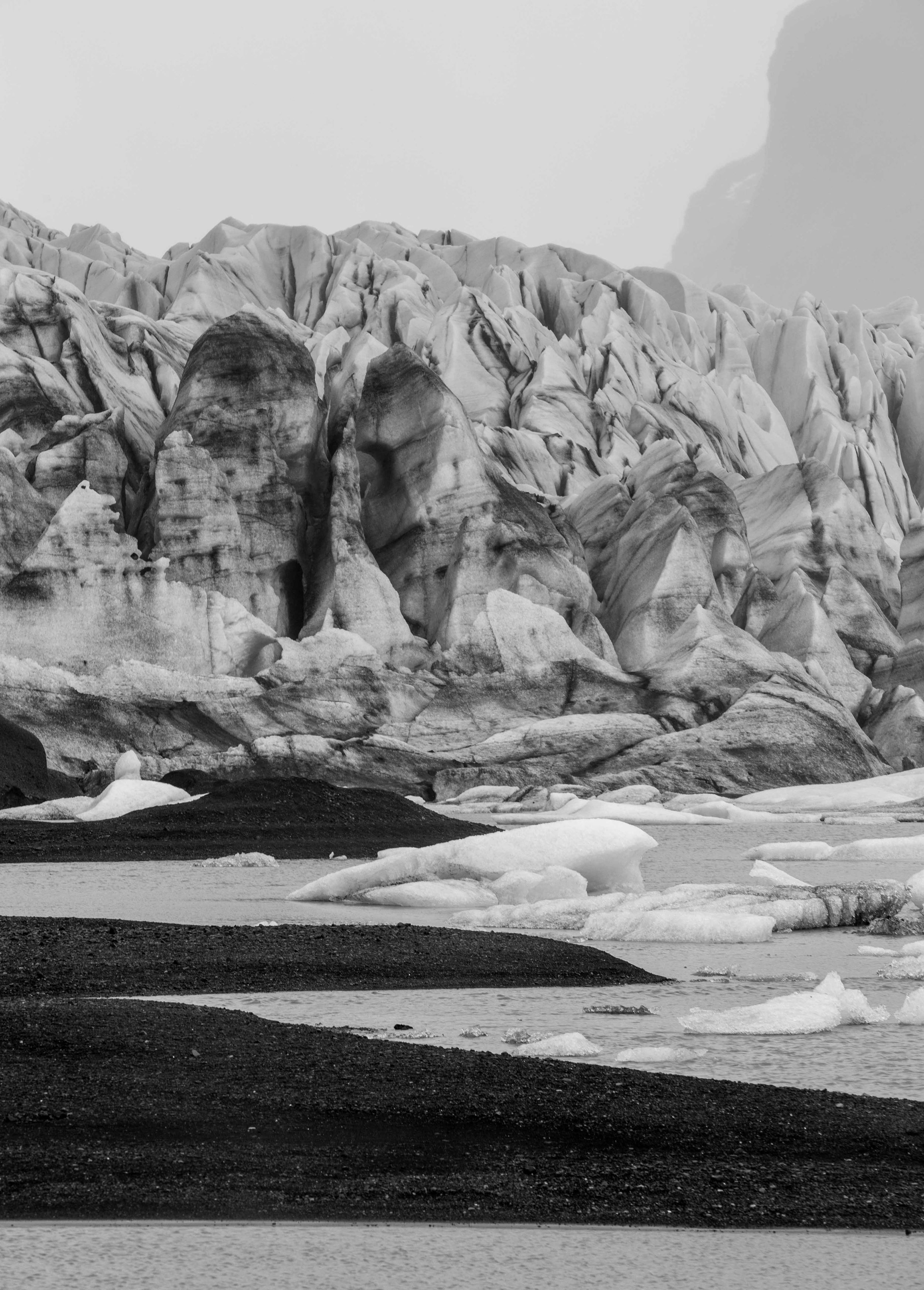 Dramatic black and white landscape of a massive glacier with jagged ice formations against a dark shoreline.
