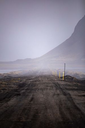 Misty rural road stretches into the distance with yellow markers and mountain silhouette in the background.