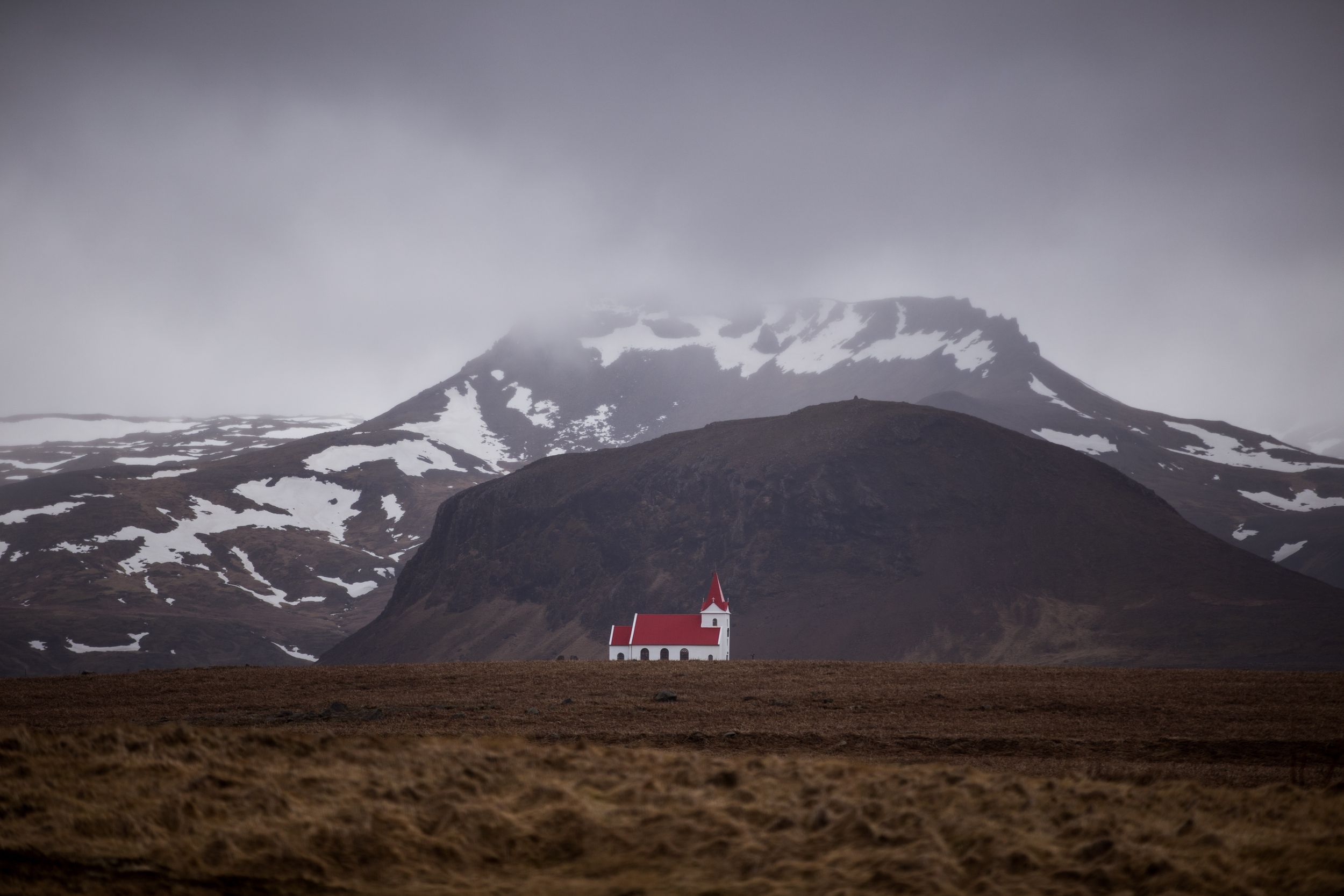 Small red church stands alone against dramatic mountain backdrop in moody Icelandic landscape.