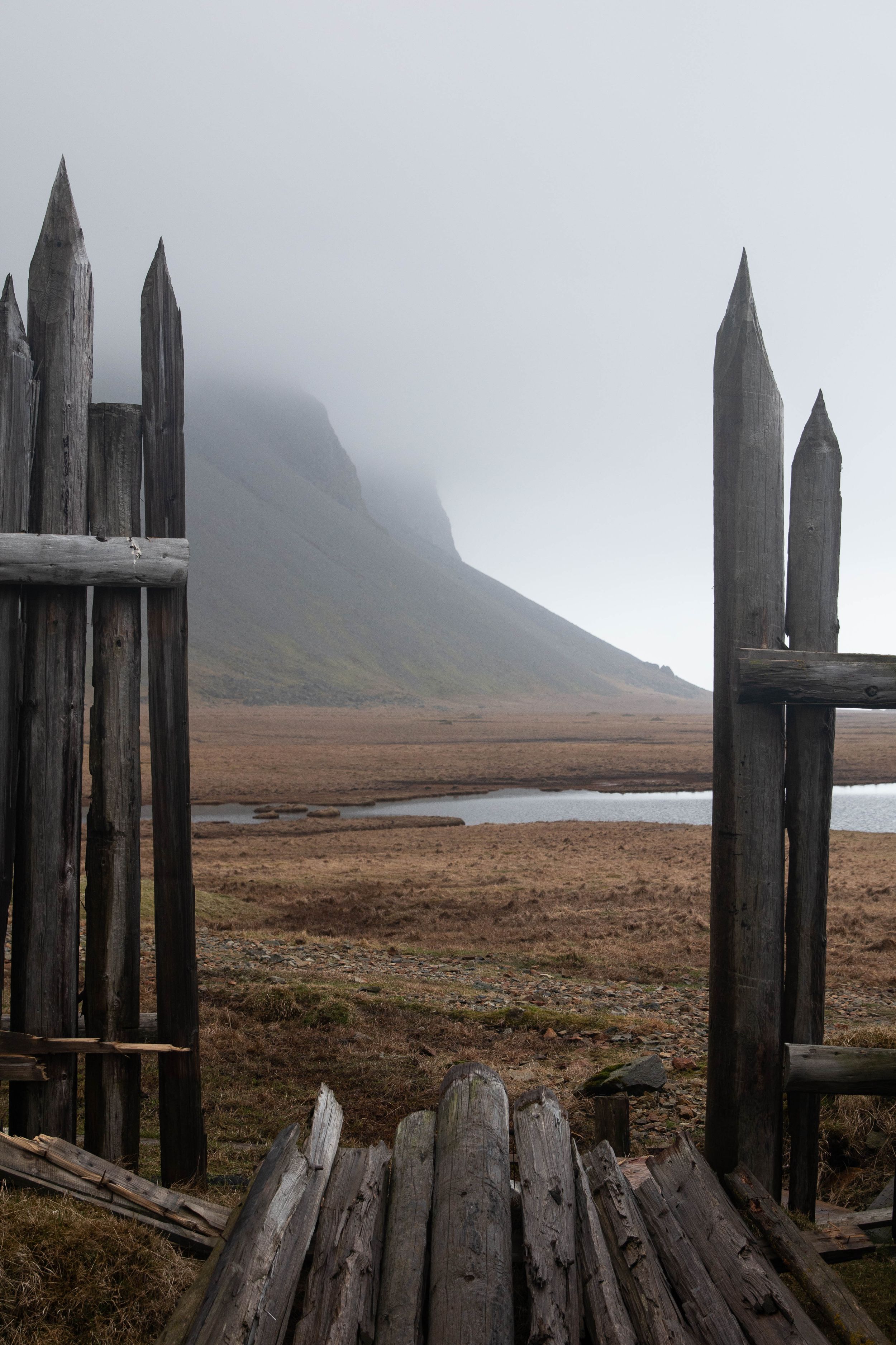 Wooden fence gate frames misty mountain view across barren landscape with small pond.