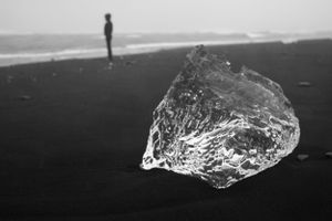 Clear ice chunk on black sand beach with silhouetted figure in the distance.