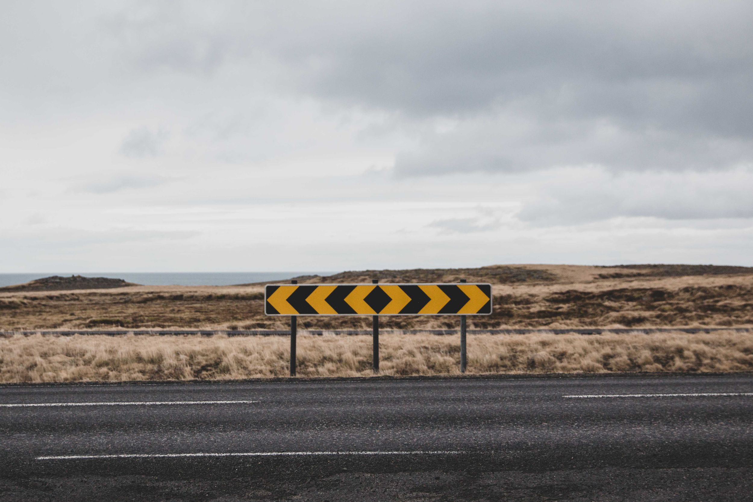 Yellow chevron warning sign on road with barren landscape and cloudy sky in background.