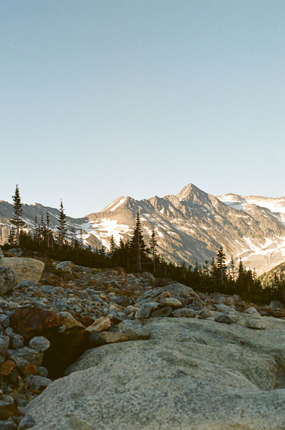 Rugged mountain peaks with snow patches rise above rocky terrain and evergreen trees at sunset.