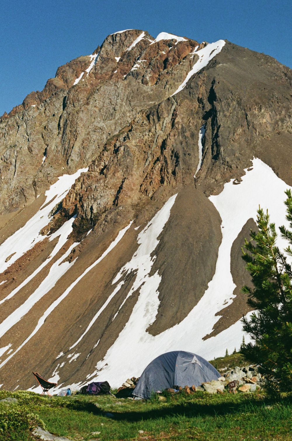 Dramatic rocky mountain face with snow patches and a small tent visible at the base.