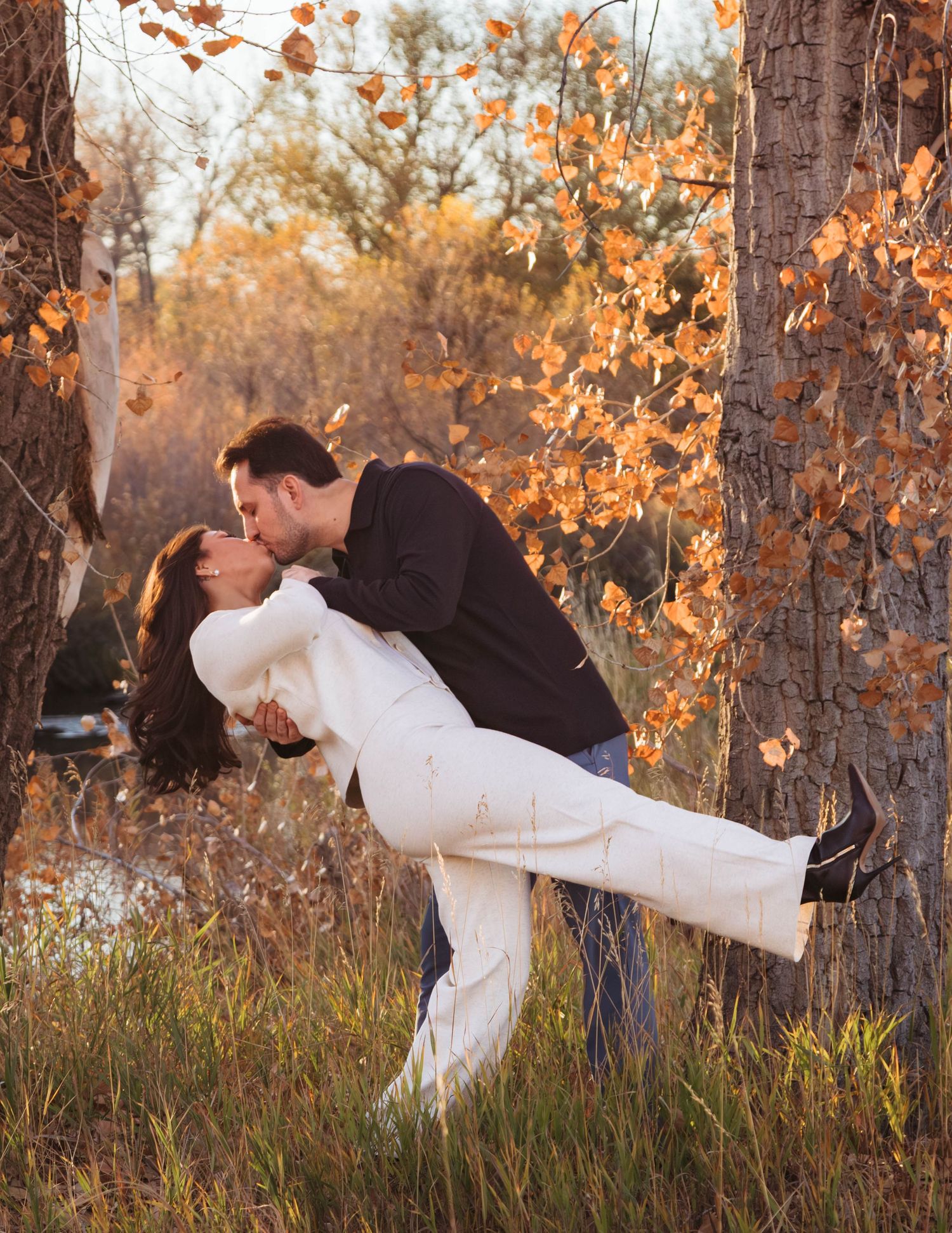 A couple shares a romantic dip kiss pose among autumn trees with golden foliage at sunset.