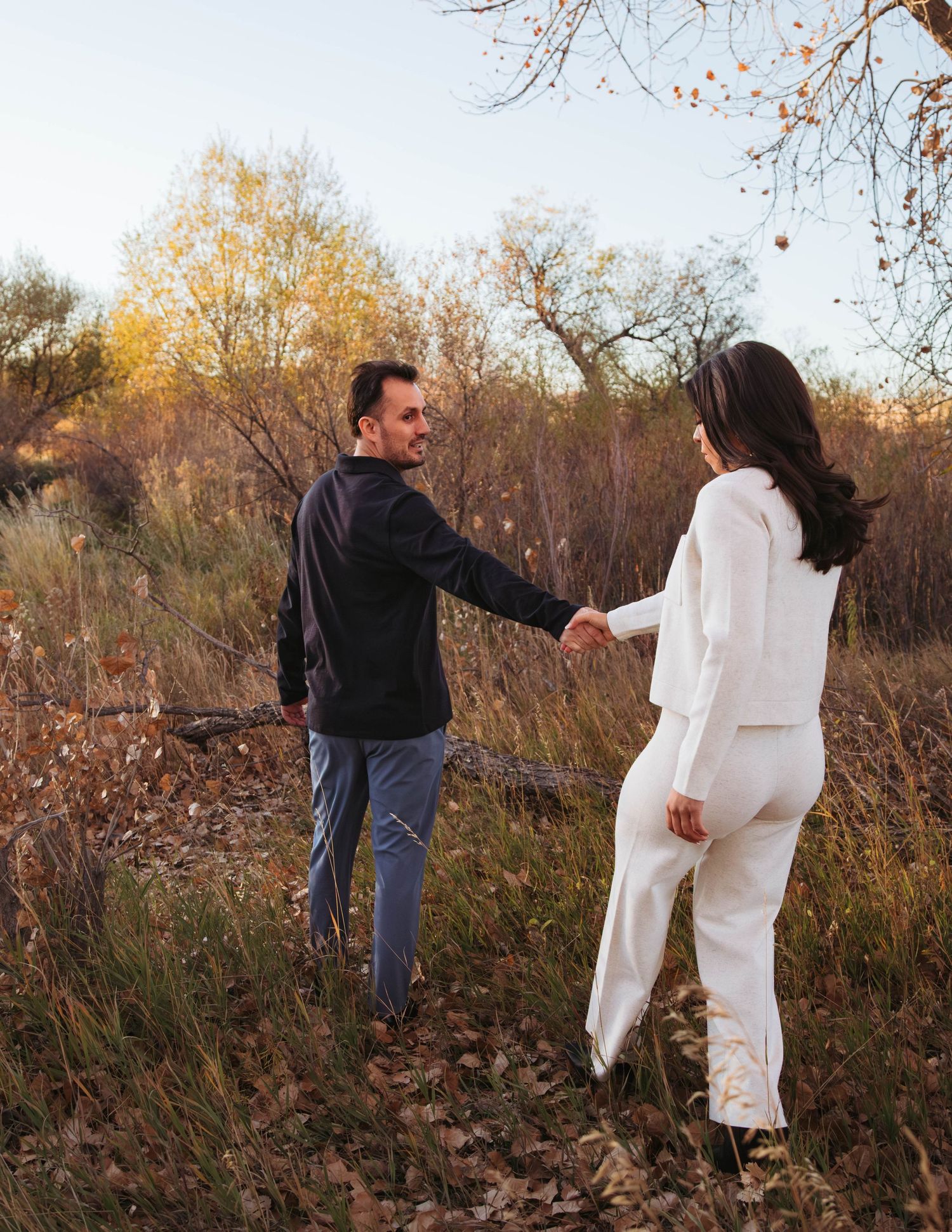 A couple in autumn foliage walks through tall grass and brush during a golden hour outdoor photo session.