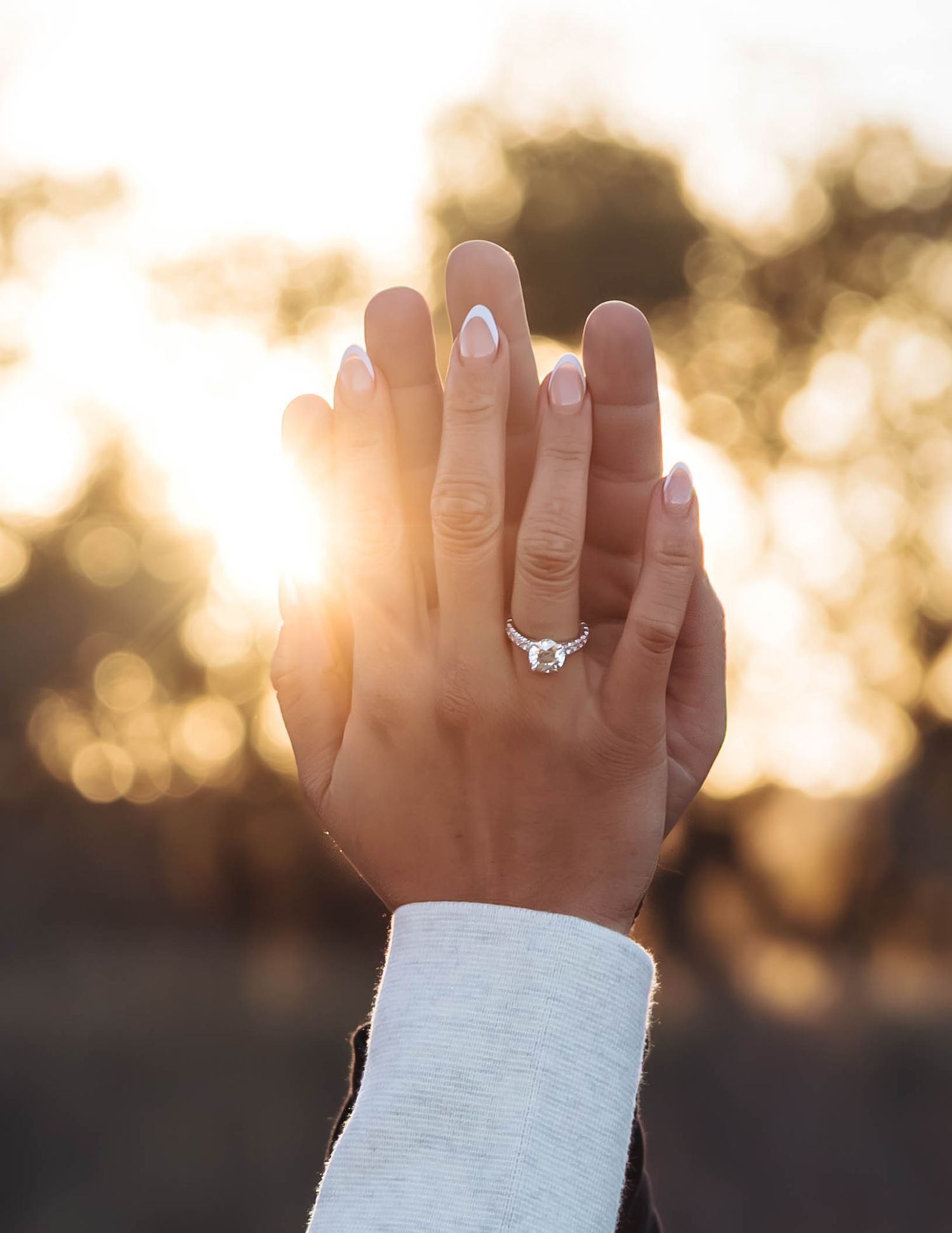 Hand wearing a sparkling diamond engagement ring held up against a warm sunset with bokeh background effect.