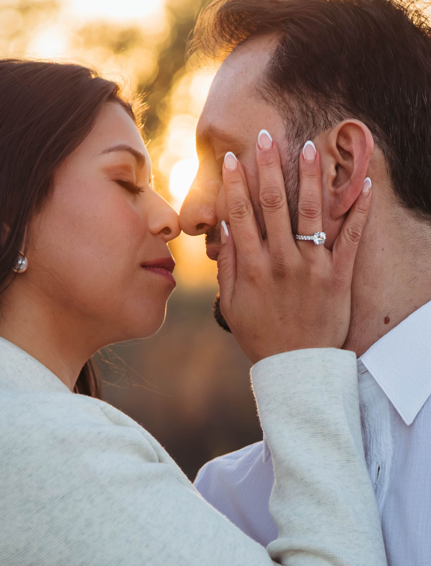 Romantic couple sharing an intimate moment at sunset with hands tenderly touching faces and engagement rings visible.
