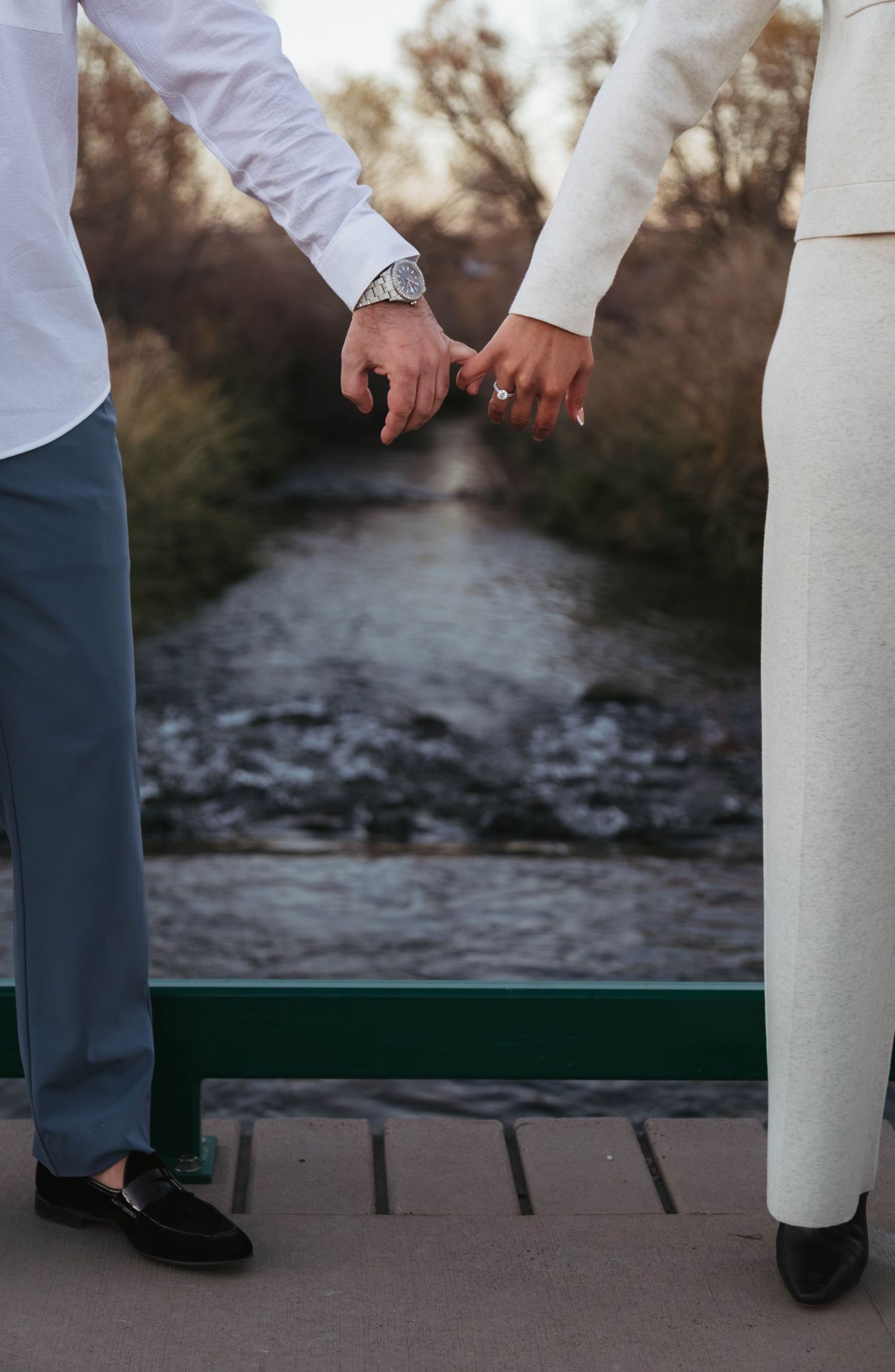 Feet in formal shoes stand on a wooden deck overlooking a scenic water view at sunset.