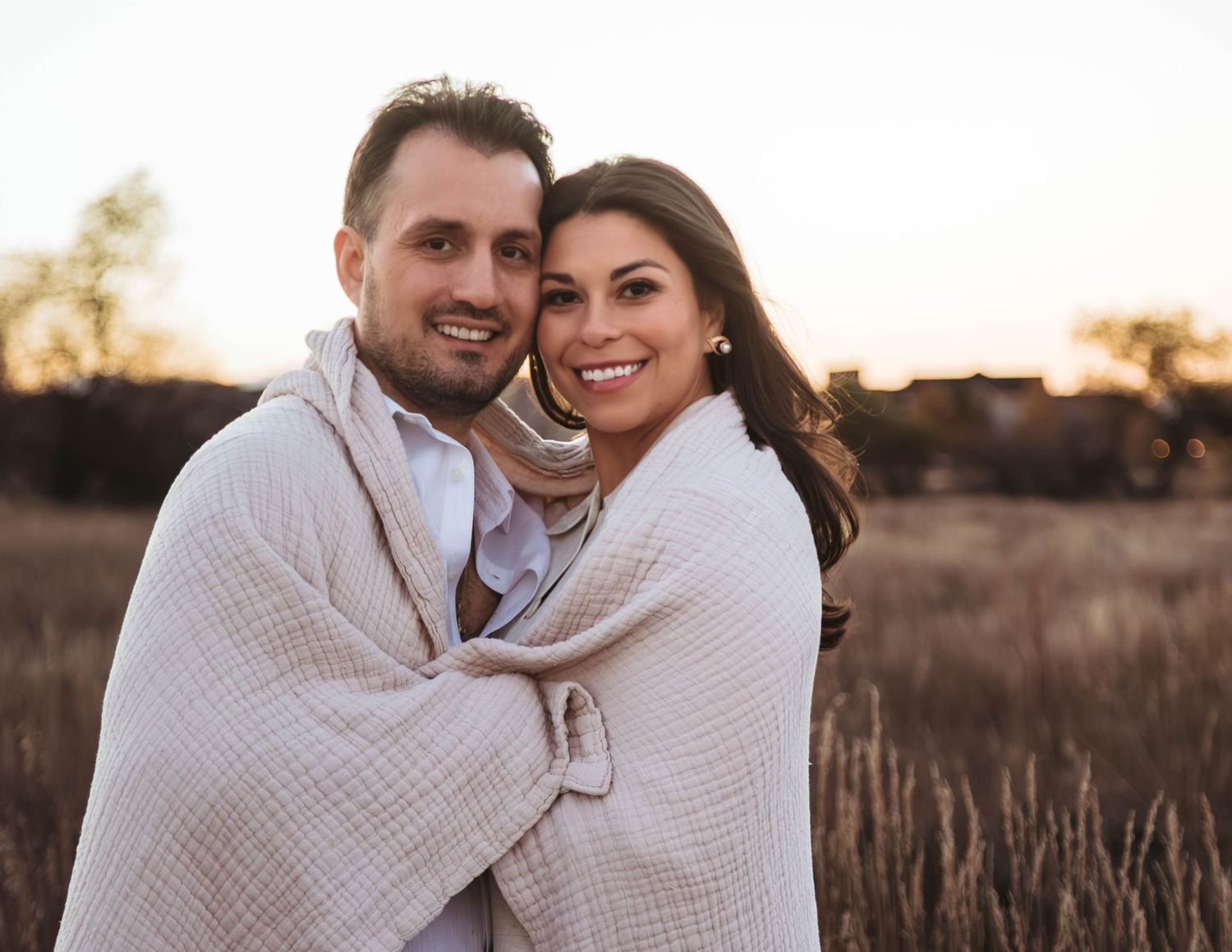 Couple wrapped in cozy white blanket embracing during outdoor engagement photo session at sunset.