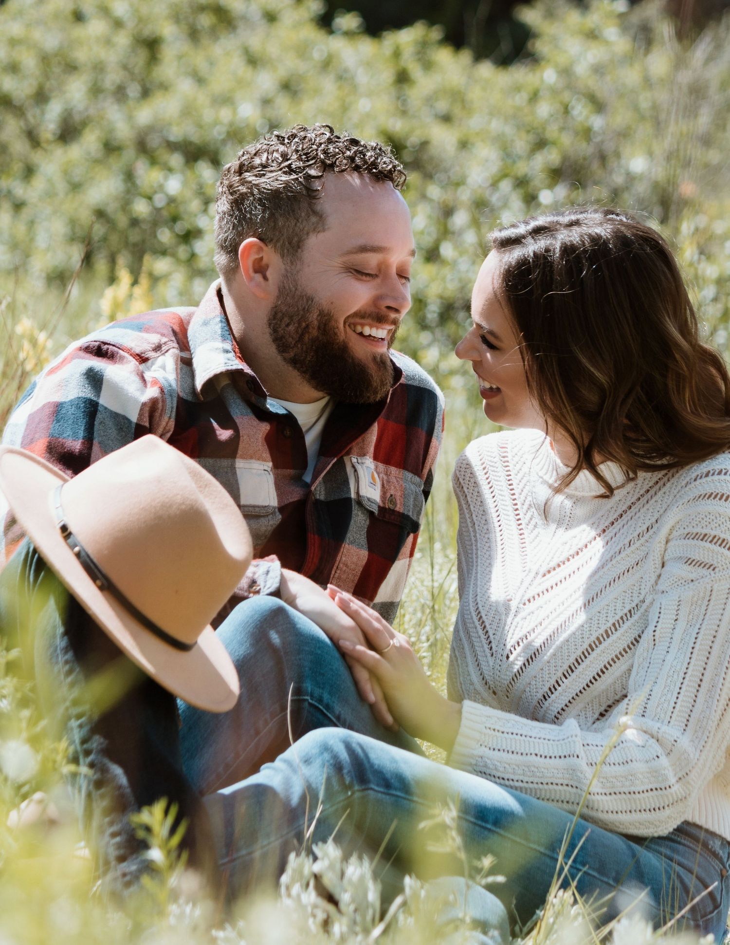 A couple shares a joyful moment while sitting in a meadow with a cowboy hat nearby.